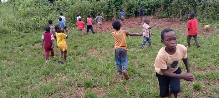 The scene depicts approximately thirteen children engaged in outdoor activity in Ngalula, Democratic Republic of the Congo. The children, appearing to be boys ranging in age from approximately 5 to 12 years, are distributed across a field of sparse green grass and reddish-brown earth. Most are dressed in casual clothing, including t-shirts and pants or shorts; some garments exhibit visible dirt or mud stains. Footwear varies, with some children barefoot and others wearing sandals. A yellow spherical object, resembling a deflated football, is visible in the upper right background. The children are in various states of movement, suggesting active play; one child in the foreground, wearing a light brown t-shirt and dark shorts/pants, is leaning forward with clenched fists, indicative of running. Another child in a yellow t-shirt and blue pants is seen from behind, moving towards the background, with significant mud staining on his lower back and pants. The background consists of dense green foliage, suggesting an undeveloped or rural landscape beyond the playing area.