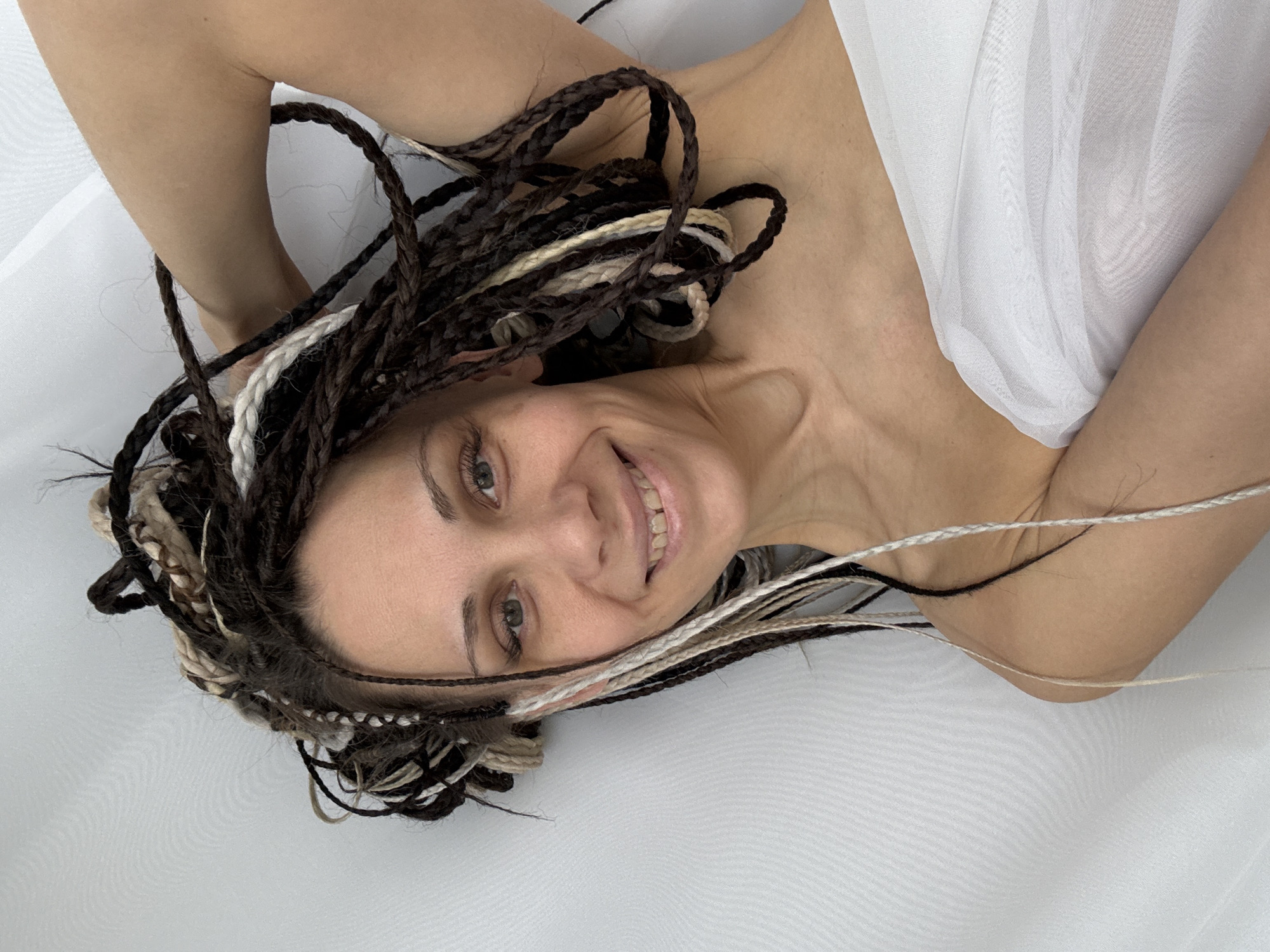 A woman with dark and light-colored braided hair is lying on a white surface. Part of a white, translucent fabric is draped over her body. The photograph appears to have been taken from above. The location is not discernible from the image.