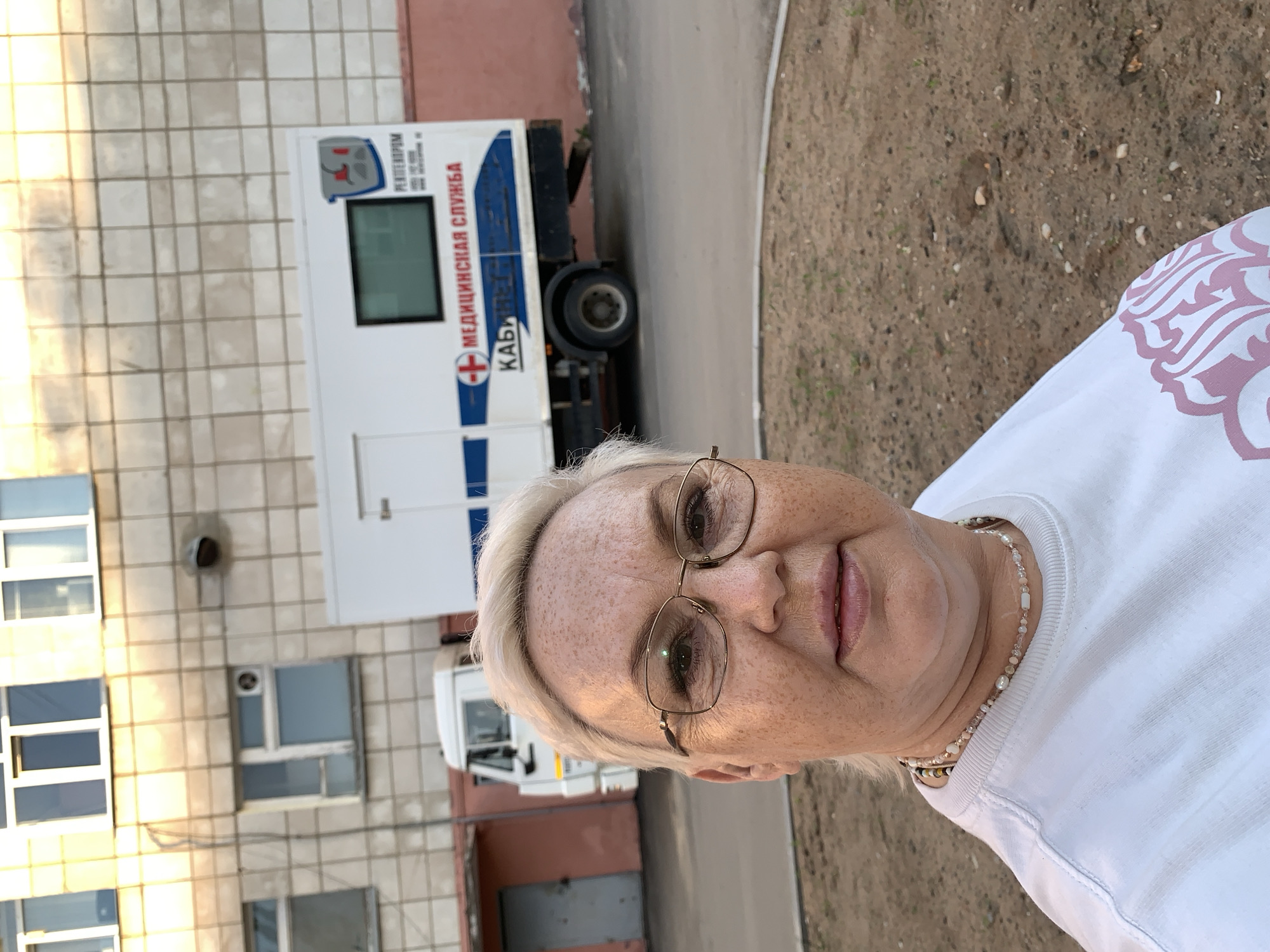 A middle-aged woman with blonde hair and glasses is the central focus, positioned in front of a building and a white truck labeled "Медицинская Служба" (Medical Service) with a red cross. The building has a light-colored facade with multiple windows. The ground is covered with dirt and some vegetation. The overall location appears to be in an urban environment, possibly in Chaikovsky, Russia, based on the Cyrillic text on the truck.