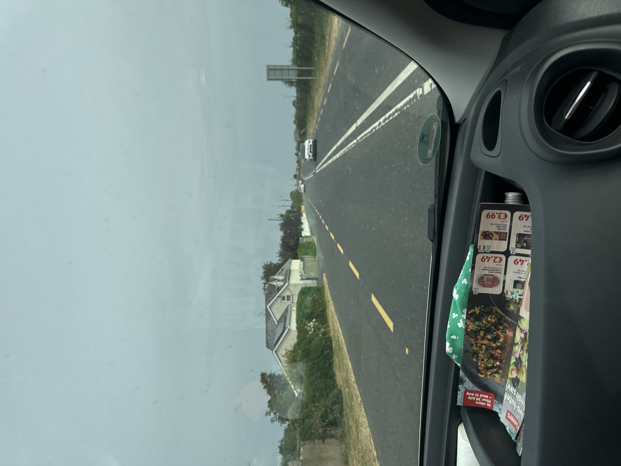 The image is taken from inside a vehicle, looking out through the windshield at a rural road in Killcommon, Ireland. The sky is overcast and a light grey. The road is paved and has yellow dashed lines marking the lanes. There are several white cars visible on the road, some in the distance and one approaching. On the right side of the road, there is a sign and dense green vegetation. On the left side of the road, there is a house with a green hedge in front of it. In the foreground, inside the vehicle, there is a storage compartment on the dashboard containing some leaflets and a green cloth item.