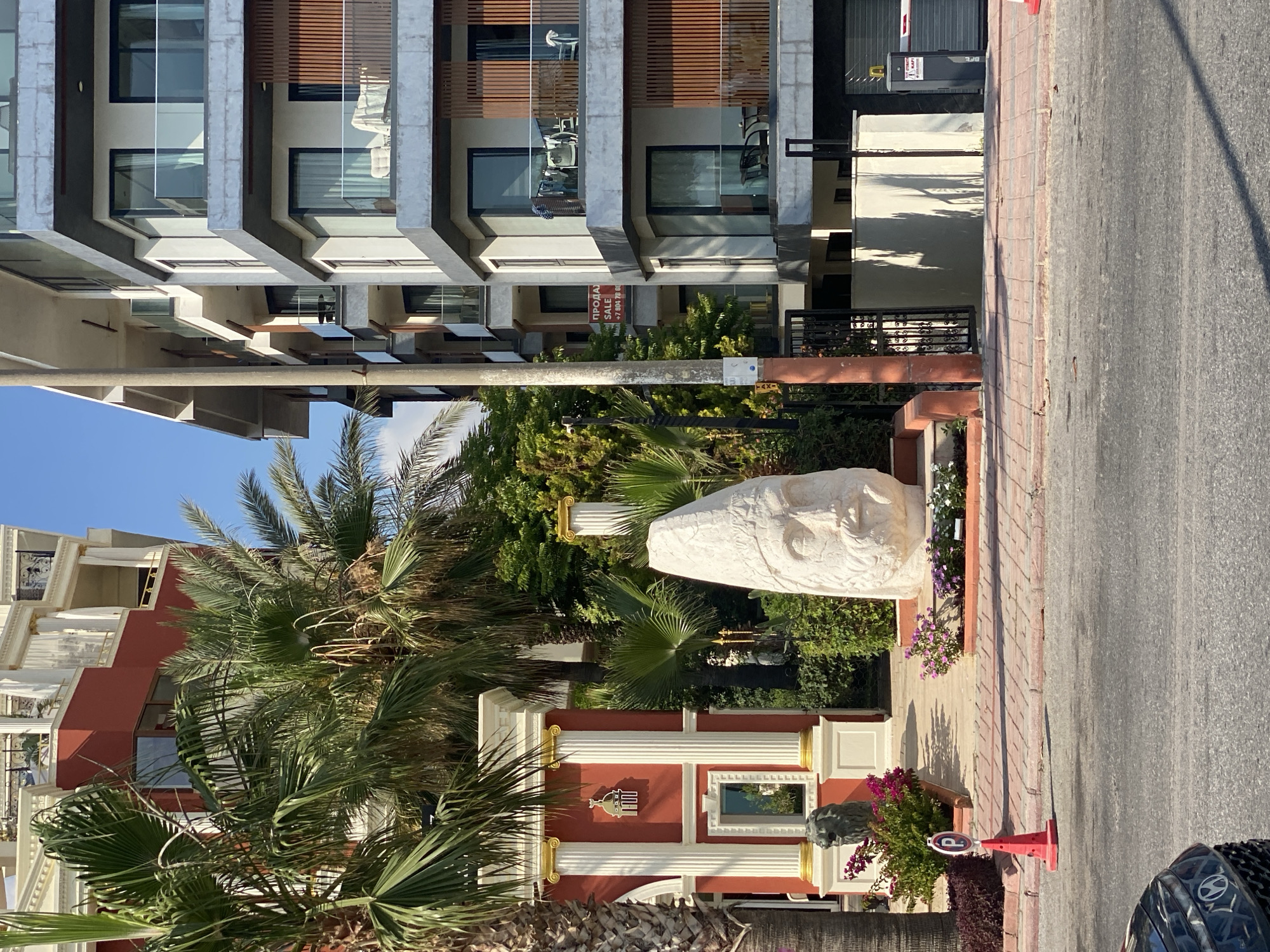 The image captures a sunny daytime scene in Alanya, Turkey, showcasing a blend of architectural styles and outdoor elements. In the foreground, a paved walkway with red and beige brick patterns leads to the entrance of a property. A bright red traffic cone with a white parking sign is placed on the walkway. To the left, lush green palm trees dominate the view, with their fronds casting shadows.
Behind the palm trees, a striking red building with white columns and gold accents stands, reminiscent of classical architecture. A carved stone lion statue is positioned at the base of the building. Further back, a massive, stylized white sculpture of a human head with prominent facial features and a helmet-like headdress is a focal point. This sculpture is surrounded by greenery and smaller flowering plants.
To the right of the head sculpture, a modern, multi-story apartment building features balconies with glass railings and wooden accents. A sign indicating "FOR SALE" in Russian and English is visible on the building. The street in front of the property is asphalted, with a dark-colored car partially visible in the lower left corner. The sky above is a clear, bright blue. The overall impression is a vibrant and eclectic streetscape, harmoniously combining historical influences with contemporary design in a Mediterranean setting.
