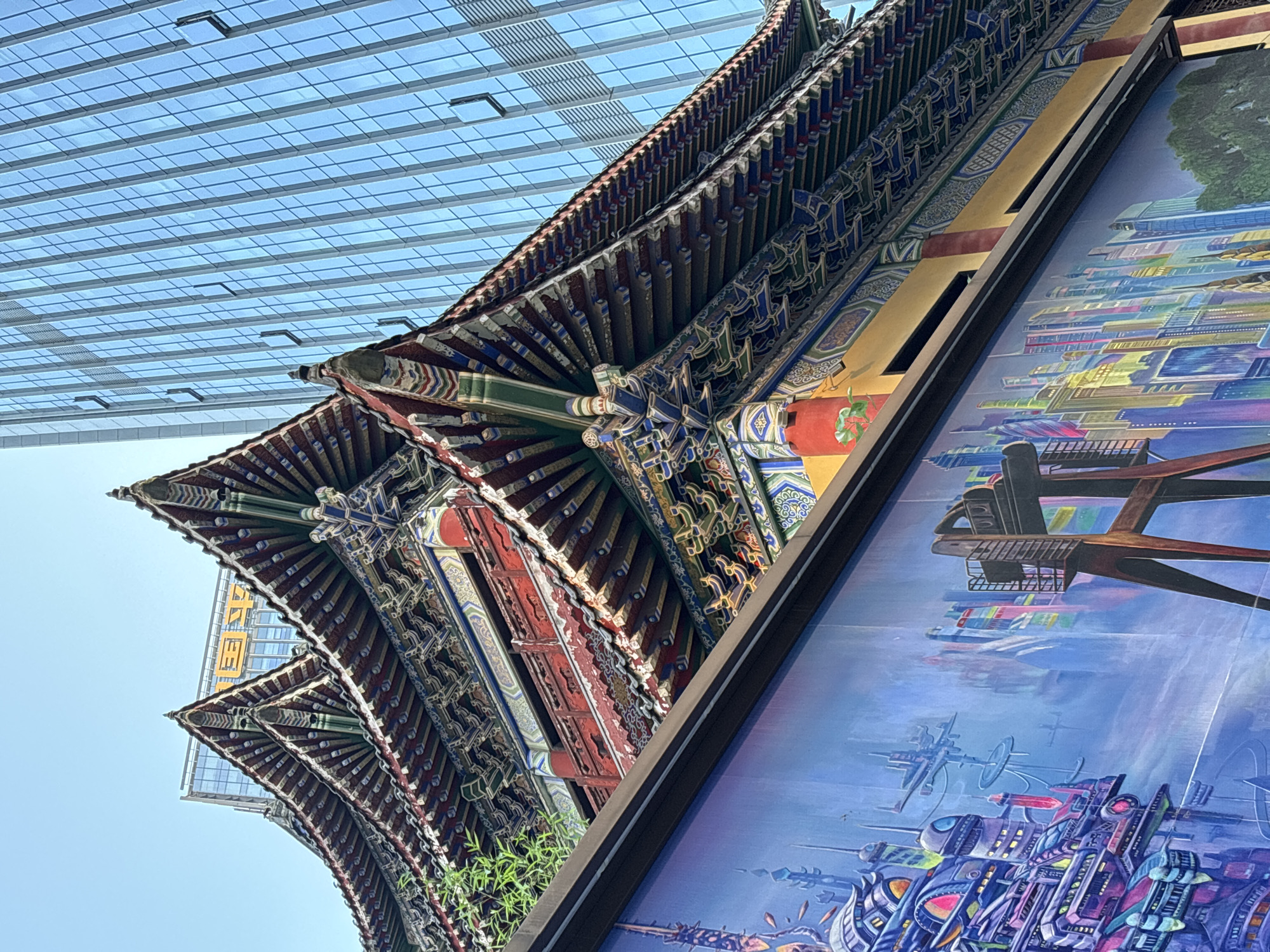 The image captures a juxtaposition of traditional and modern architecture in the Yuzhong District of China. An ornate, multi-tiered traditional Chinese building with intricate carvings and vibrant colors is viewed from below. The building's roof features elaborate bracketing and detailed painting, showcasing the craftsmanship of traditional Chinese architecture. In the background, a modern glass building rises, its grid-like facade reflecting the sky. To the right of the traditional building is a painting of a futuristic cityscape. The architectural contrast highlights the blending of historical heritage and contemporary development in this urban setting.