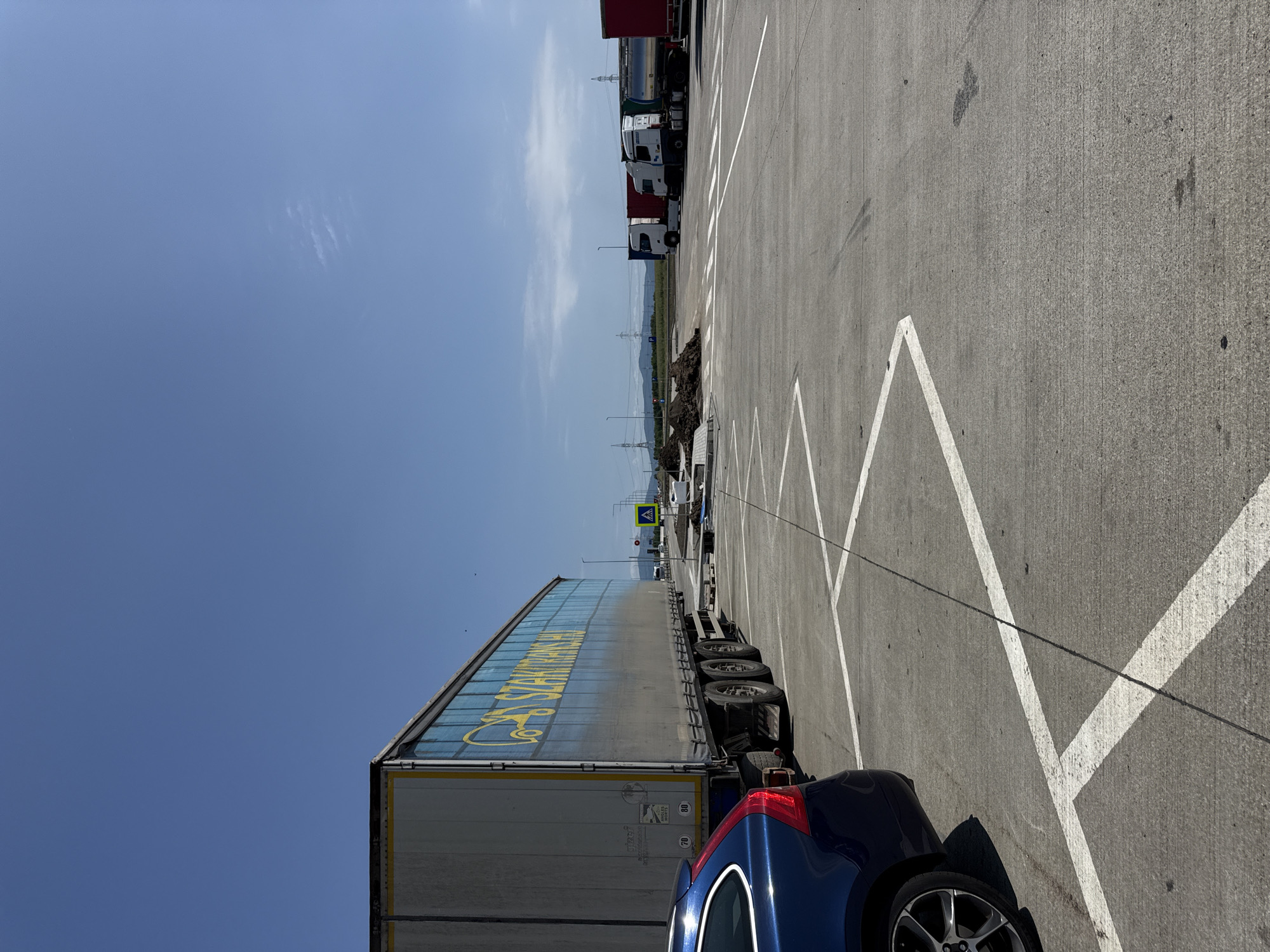 In Sanpetru, Romania, a blue Volvo car is parked in a designated spot marked by white lines on a paved lot. To the left, a large semi-trailer truck is parked, its side displaying the logo "SZAKTRANS.HU". In the background, several other semi-trailer trucks are lined up, some white and some red. A pile of dark soil is visible in the middle ground. A pedestrian crossing sign is also present. The sky is clear and blue.
