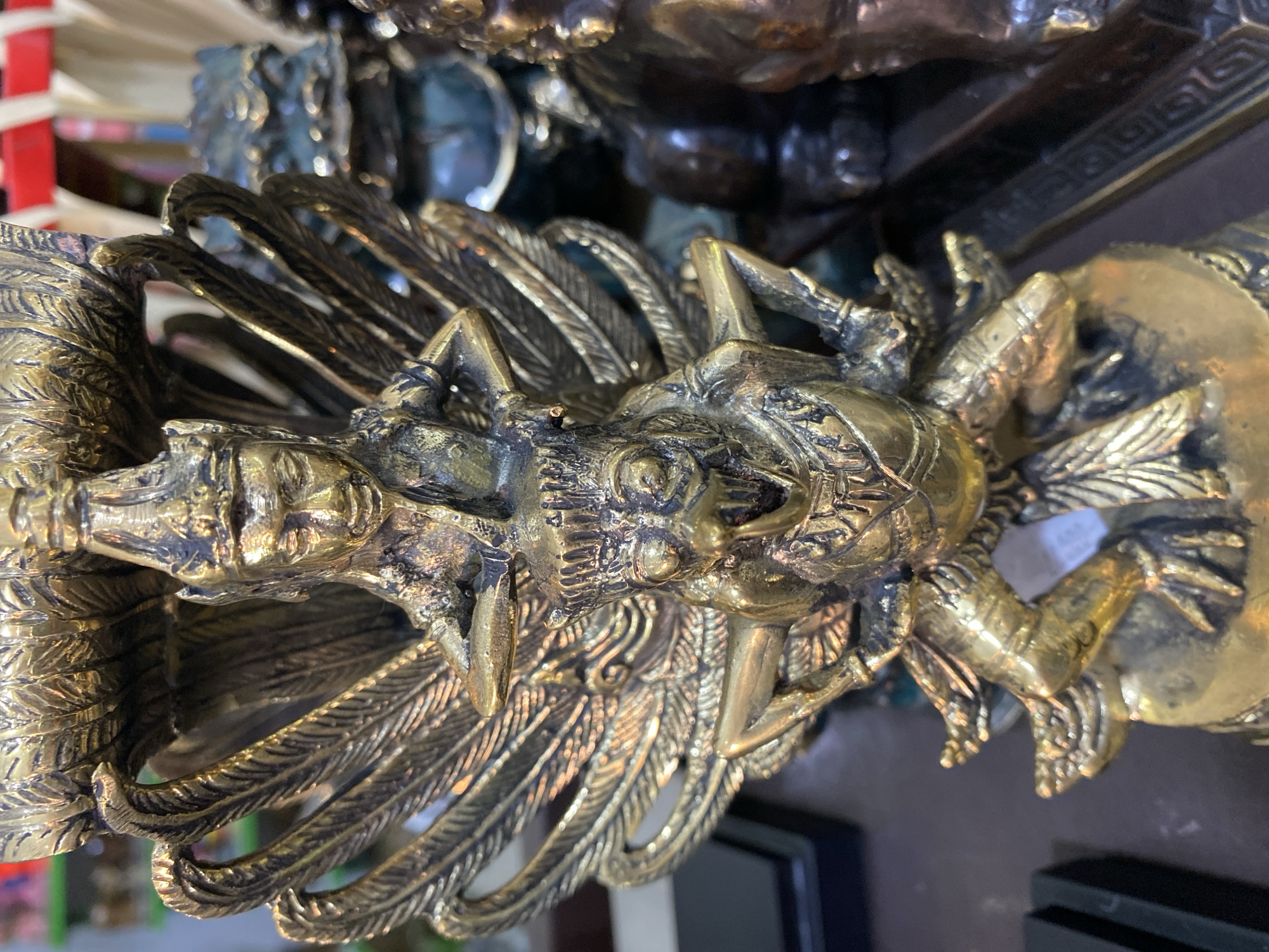 The image depicts a close-up view of a golden, intricately detailed statue of a dragon-like creature with feathers, located in Nusa Dua, Indonesia. The statue appears to be made of metal, possibly brass or bronze, and is positioned at an angle, capturing the head and upper body of the creature. The surrounding environment is blurred, suggesting an indoor setting.