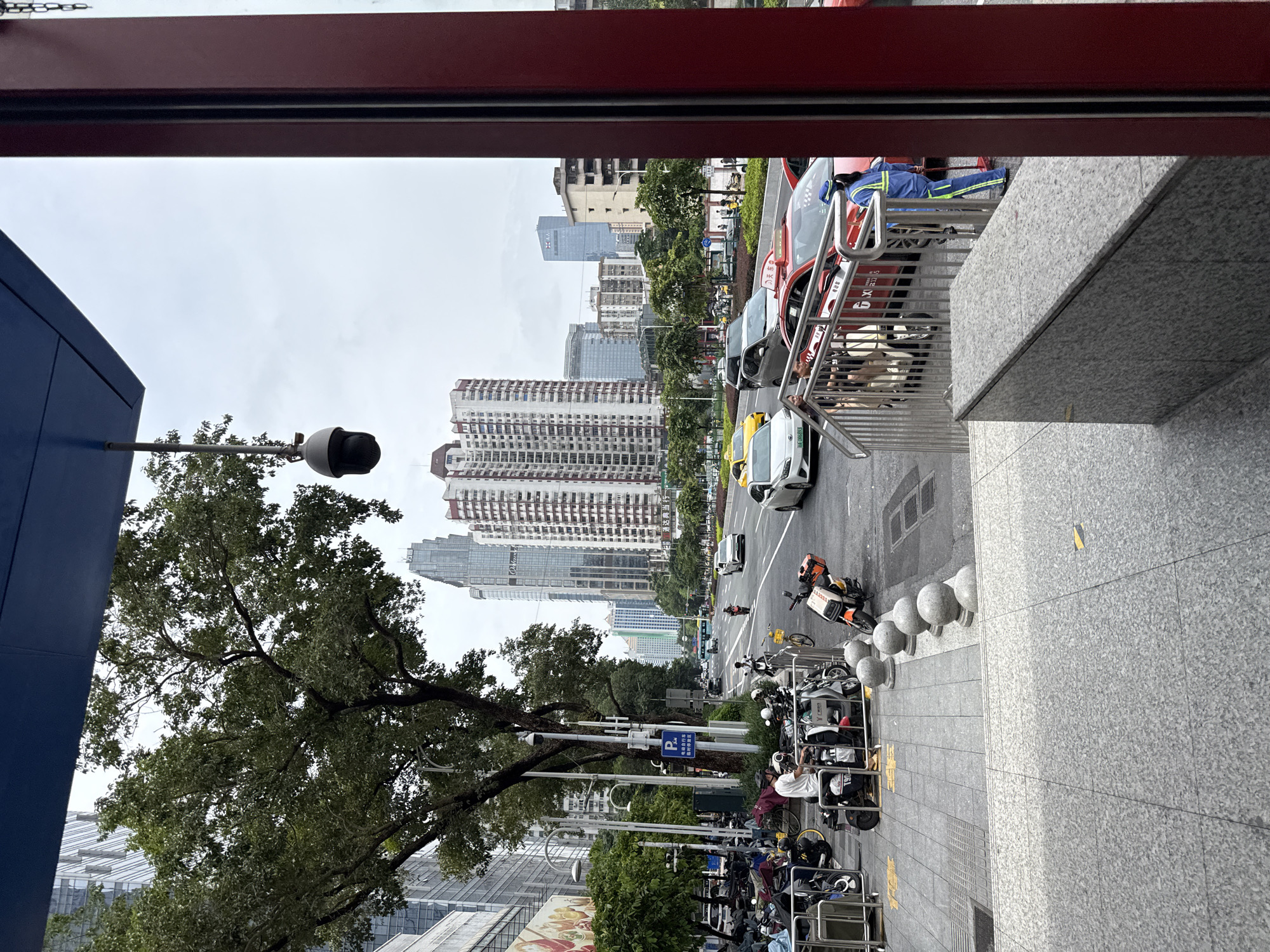 The image captures a street scene in Tianhe District, China, on an overcast day. A sidewalk with a gray, speckled pavement is in the foreground, leading up to a building with a red facade on the right. A set of metal stairs with a railing goes up from the sidewalk. To the left of the stairs, a row of parked motorcycles and scooters is visible, some covered. A person in a white t-shirt is standing among the motorcycles. Beyond the sidewalk, a road with multiple lanes of traffic is present. Several cars, including yellow taxis and a red car with Chinese characters and a driver in a blue uniform, are driving on the road. A person in a blue uniform with bright green reflective stripes is sweeping the street near the red car. Trees line the street, and modern high-rise buildings dominate the background, indicating an urban environment. A security camera is mounted on a pole near the trees. The overall atmosphere is that of a busy city street.