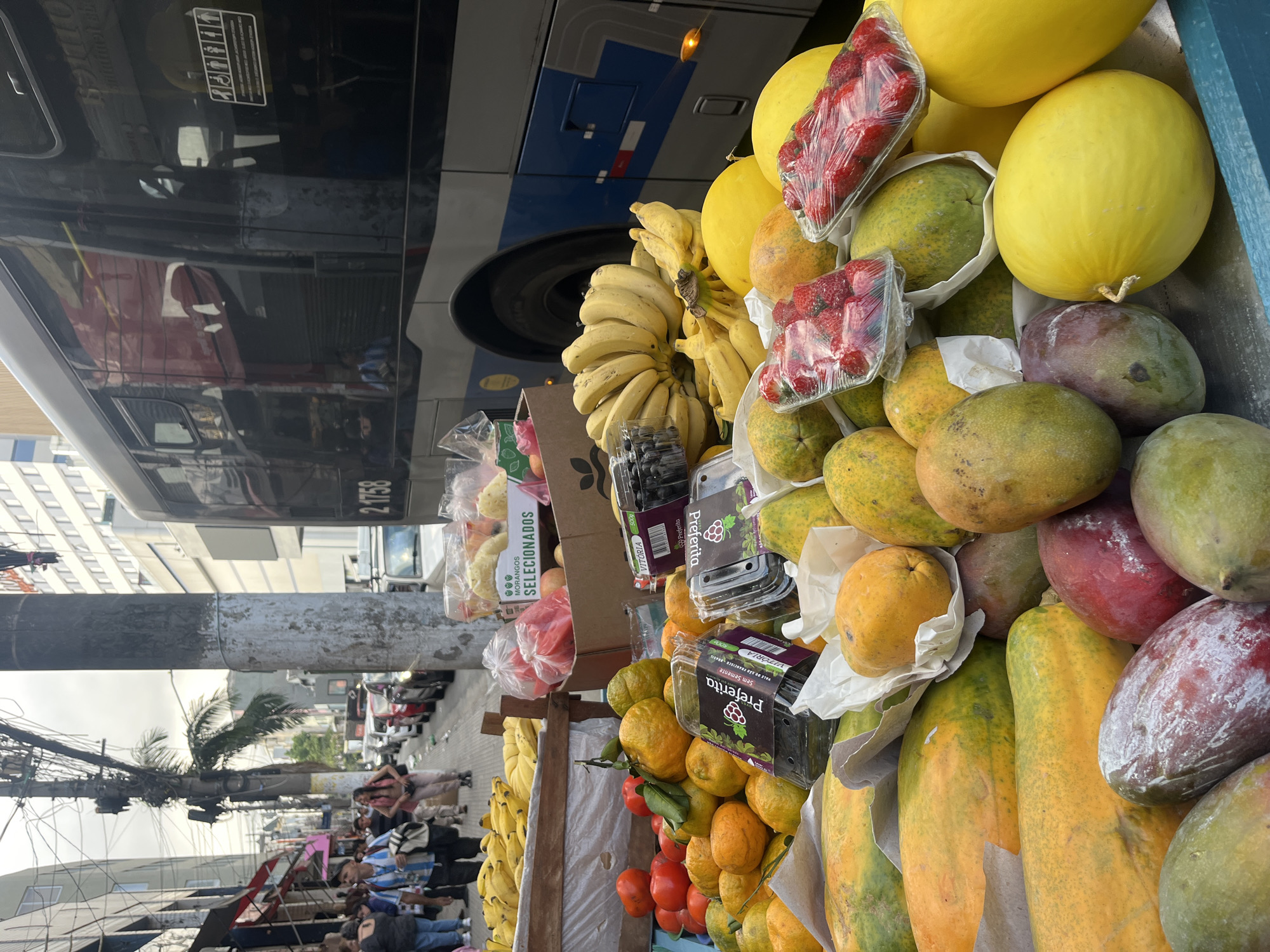 The image depicts a street scene with a fruit stand in Unknown, Unknown. The fruit stand is located next to a bus, and the stand is stocked with bananas, yellow melons, strawberries in plastic containers, mangoes, papayas, oranges, tomatoes, and boxes of what appear to be blueberries or blackberries. People are seen walking on the sidewalk in the background. The street is lined with buildings, and overhead power lines are visible.