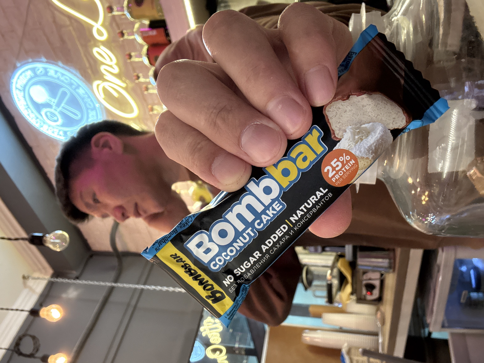 A hand holds a "Bombbar Coconut Cake" protein bar. The packaging states "No Sugar Added | Natural" in both English and Russian. The bar is chocolate covered and has a white filling. The text "25% Protein per serving" is visible on the packaging. In the background, a person is visible, along with a neon sign that reads "One L" and includes a logo of a spoon and fork. String lights hang overhead. A coffee station with equipment and containers can be seen further in the background. The OCR text mentions the manufacturer and the city Khimki, Russia.