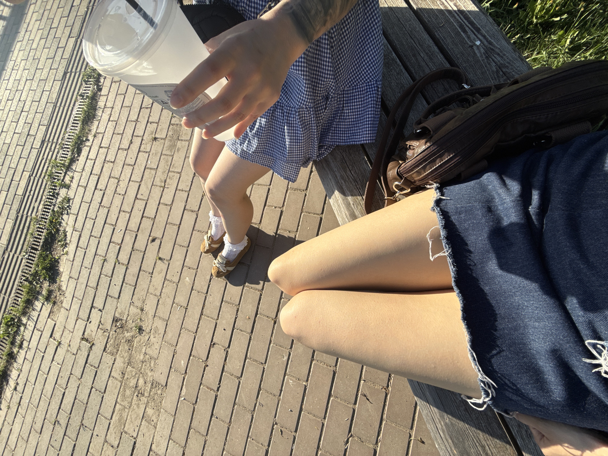 The image, captured from a high-angle perspective, shows two people seated on a wooden bench. The person on the left is holding a plastic cup with a straw, wearing a blue and white gingham dress, white socks with frills, and brown shoes. The person on the right is wearing a denim skirt. A brown satchel-style bag sits next to the person on the left. The bench sits on a brick-paved area. A grassy drainage ditch runs along the edge of the brick paving. The location is identified as being within the city of Votkinsk, Russia.