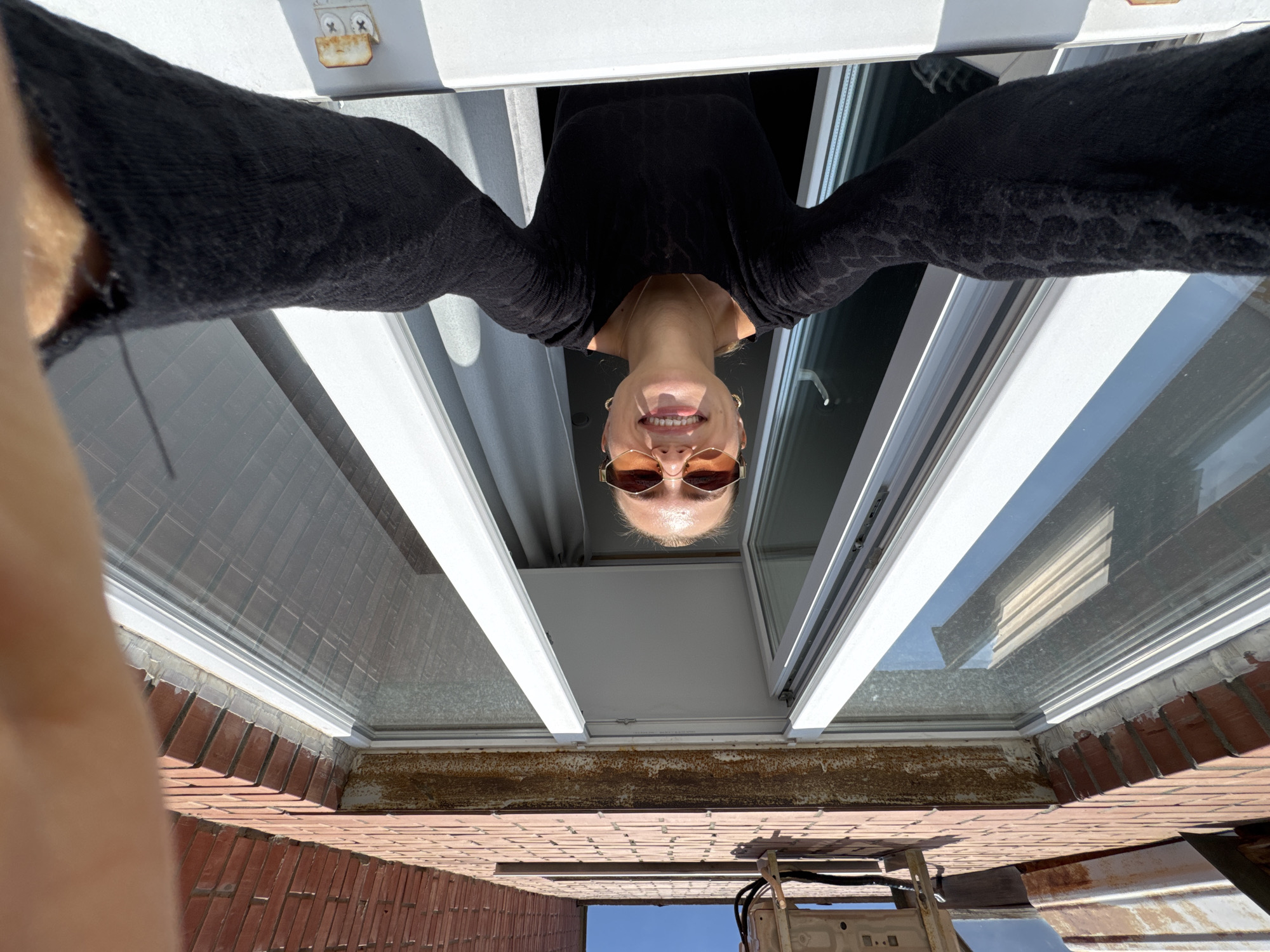 An inverted, low-angle selfie captures a person, likely a woman, smiling cheerfully while looking downwards from an open window or skylight. Her face, seen upside down, is framed by sunglasses with amber-tinted lenses, and she wears a black, textured, long-sleeved top and a delicate gold-colored necklace. Her hair appears light-colored and pulled back or short.
The person's arms are outstretched, contributing to the wide-angle, almost fisheye perspective. Around her, the white frames of the window or skylight are prominent, with glass reflecting hints of the interior and sky.
Below the window structure, a weathered red brick surface is visible, possibly a wall, ledge, or roof. On this brick surface, to the lower right, there's what appears to be an outdoor unit, possibly an air conditioner or a heater, with some cables attached. Further down, a clear blue sky is visible, indicating bright daytime conditions. There is also a metal flashing or gutter with visible rust and discoloration along the edge of the brick structure. A small, rusty metal bracket with two screws is also visible attached to the white window frame in the upper left.
The image conveys a playful mood due to the subject's smile and the unusual, inverted perspective. No discernible text is visible in the media.