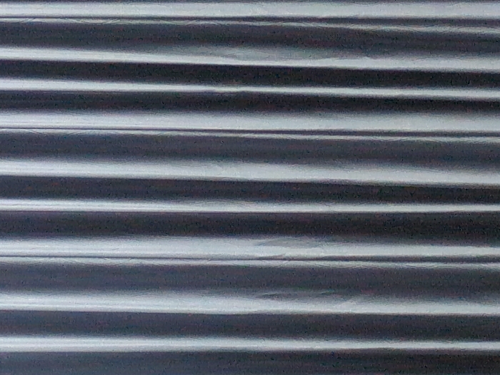 The image displays a vertically oriented, full-frame view of a textured surface, characterized by a series of parallel, vertical ridges and troughs. This creates an alternating pattern of bright, reflective highlights and darker shadowed areas, consistent with corrugated metal or ribbed fabric. The color palette is monochromatic, primarily grey tones with silvery accents. No individuals, discernible objects other than the textured material, or specific actions are visible within the frame. The visual content provides no information to identify a specific location, including any location within the city of Thành phố Đà Lạt, Vietnam.