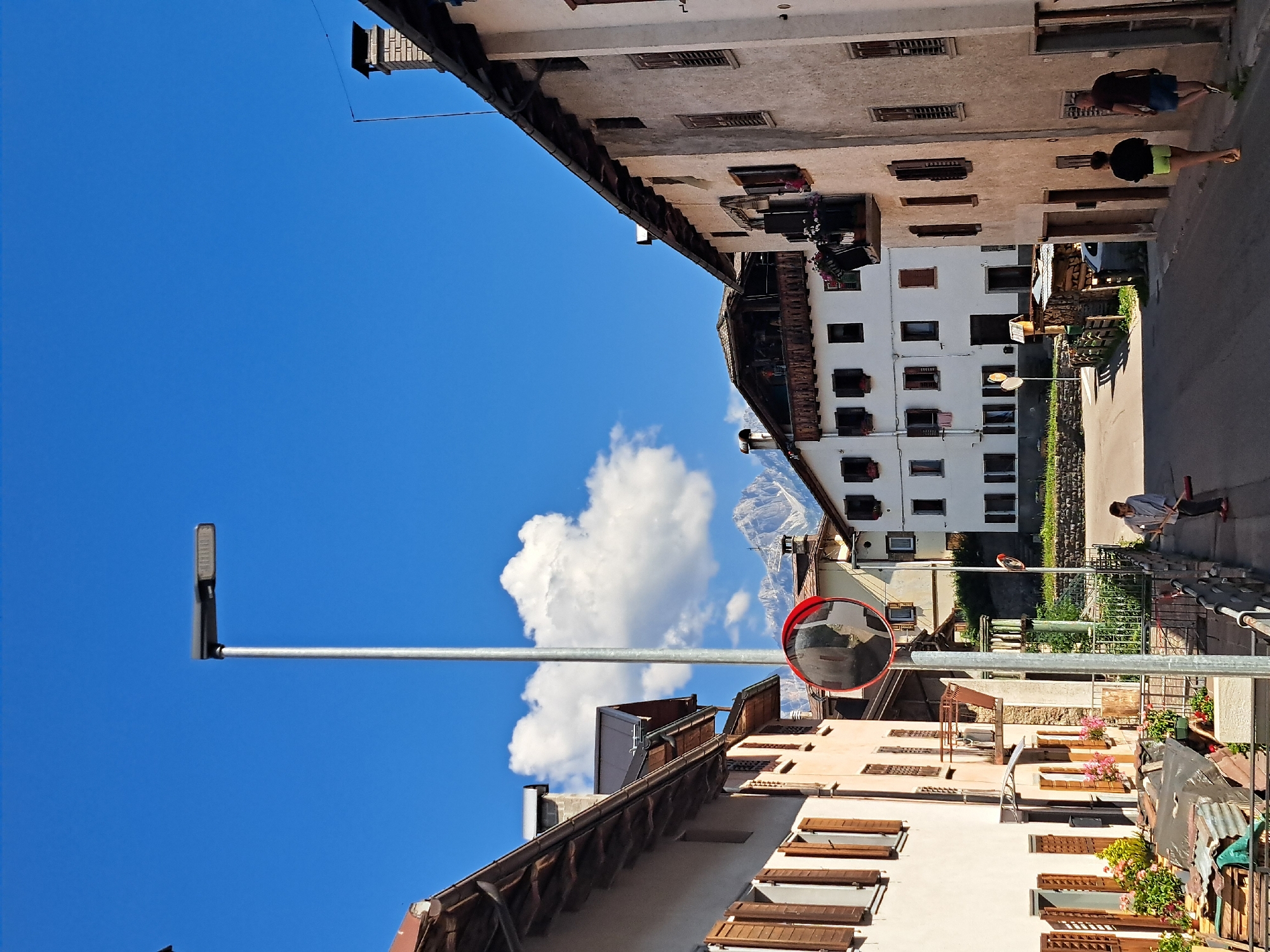 The image depicts a street scene in what appears to be a European town, possibly in the Alps due to the distant mountain visible. The buildings lining the street are multi-story structures with traditional architecture, featuring wooden shutters and balconies adorned with flowers. The sky is a vibrant blue with a single, fluffy white cloud.
There are a few people visible in the image. One person, wearing a dark t-shirt and blue shorts, is walking down the street. Another person, in a black top and neon green shorts, is also walking. Further down the street, a person is using a red broom to sweep the pavement.
Notable objects include a street lamp, a convex mirror mounted on a pole which likely serves as a traffic mirror, and various potted plants and flowers on windowsills and balconies. The buildings show signs of age and character, with weathered facades and wooden elements.
The time of day appears to be daytime, likely late morning or afternoon, given the bright sunlight casting shadows. The weather is clear and sunny. The overall impression is one of a quiet, picturesque town on a pleasant day. No visible text is present in the image. The orientation of the image is such that the bottom of the scene appears at the top and vice versa.