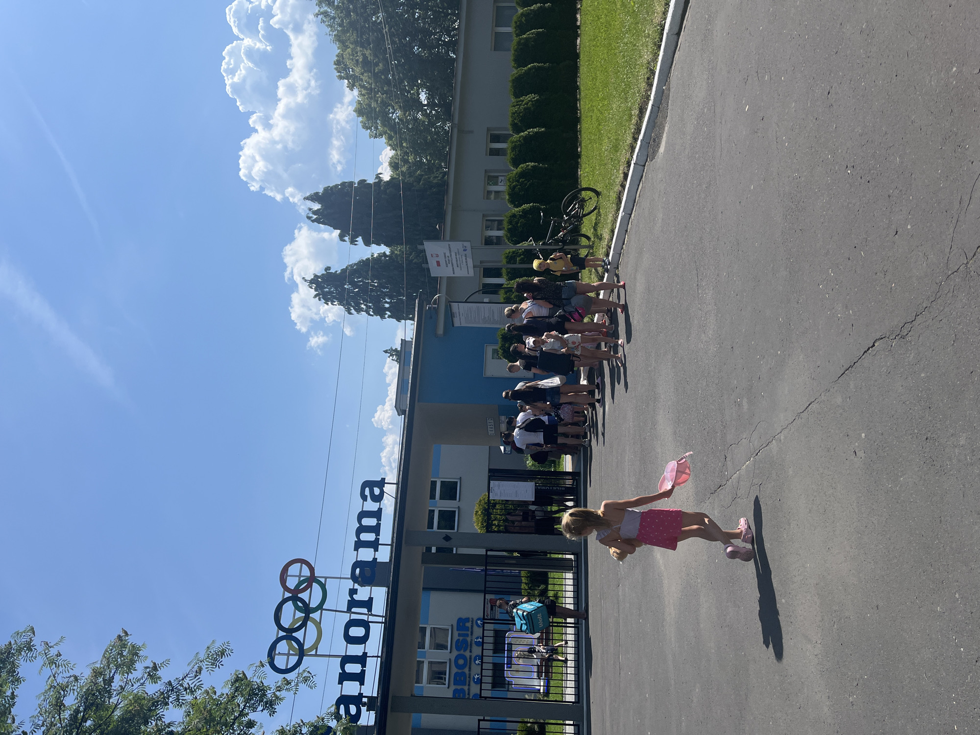 The image shows the entrance to the "Panorama" facility, which features the Olympic rings logo above the name. A line of people, including children and adults, are waiting at the entrance. Some are holding bags and some appear to be children with toys. A young girl in a pink skirt and white top is walking away from the camera, holding a pink object. A bicycle is parked to the right of the entrance. The environment is sunny with a blue sky and scattered clouds. The facility appears to be a recreational or sports complex.