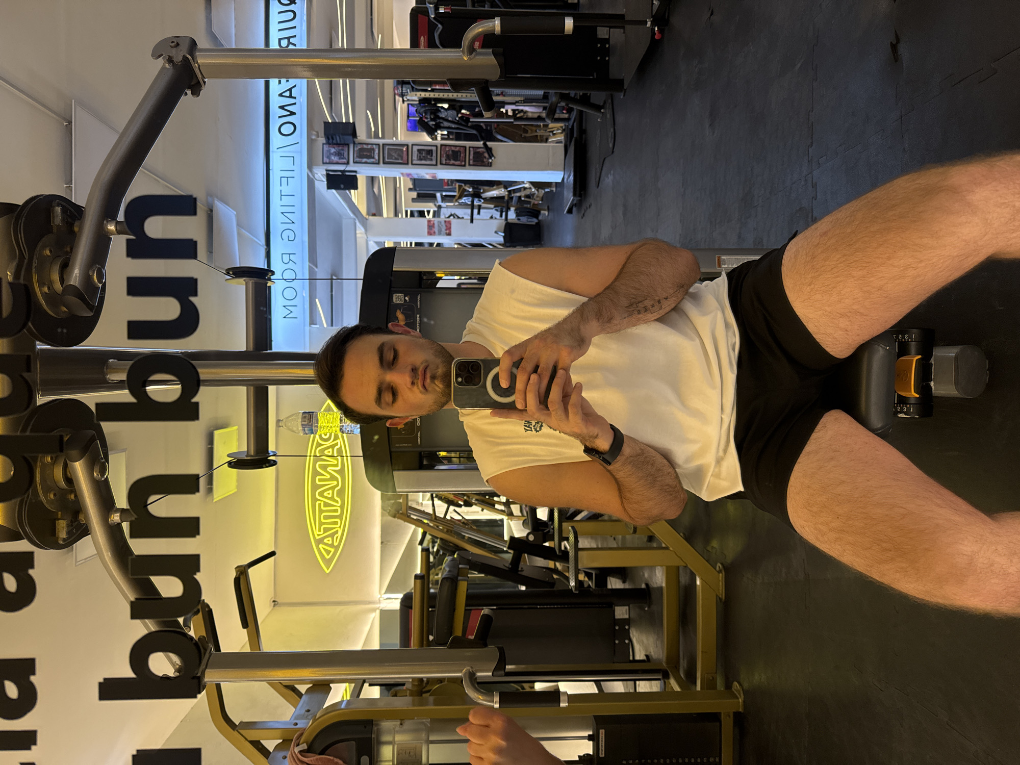 A man is taking a selfie in a gym. He is wearing a white tank top and black shorts. The gym has various exercise machines. A neon sign reads "TATAMAG," and a sign above it reads "WEIGHTLIFTING ROOM." The location is likely in Puebla, Mexico.