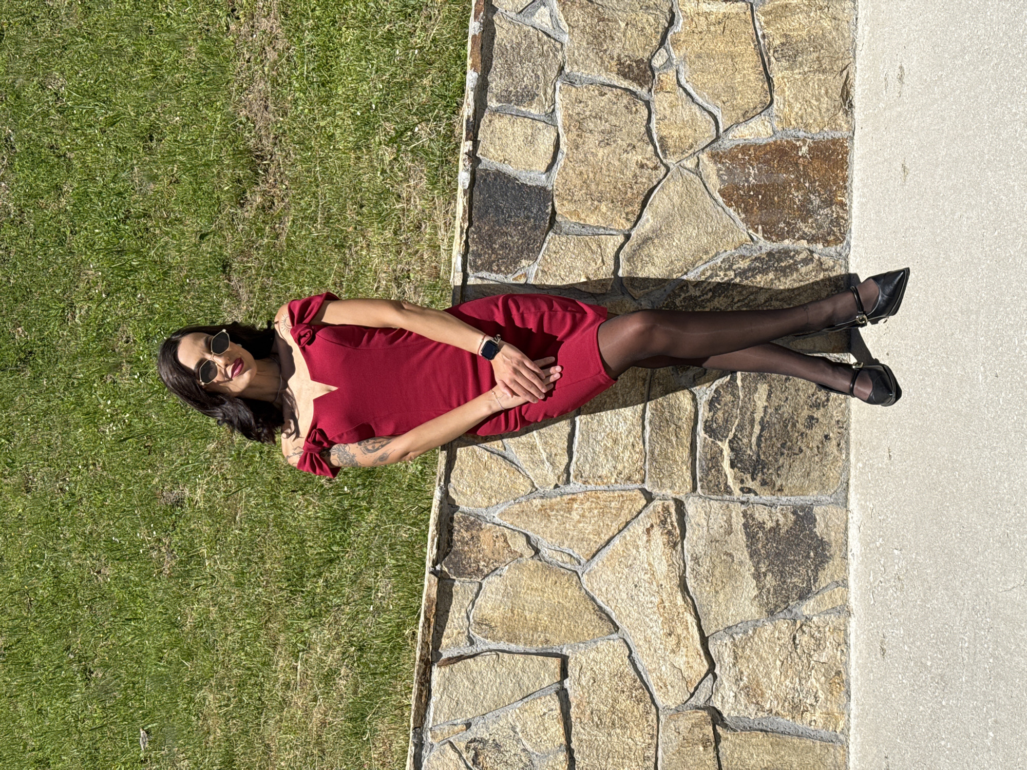 A woman in a maroon dress, black tights, and black heels lies on a stone pathway. She is wearing sunglasses and a smartwatch. The pathway is bordered by green grass on one side and a concrete slab on the other. The location is likely outdoors, potentially in a park-like setting or residential area in Ribaritsa, Bulgaria. The image appears to be taken during the day with good lighting conditions.