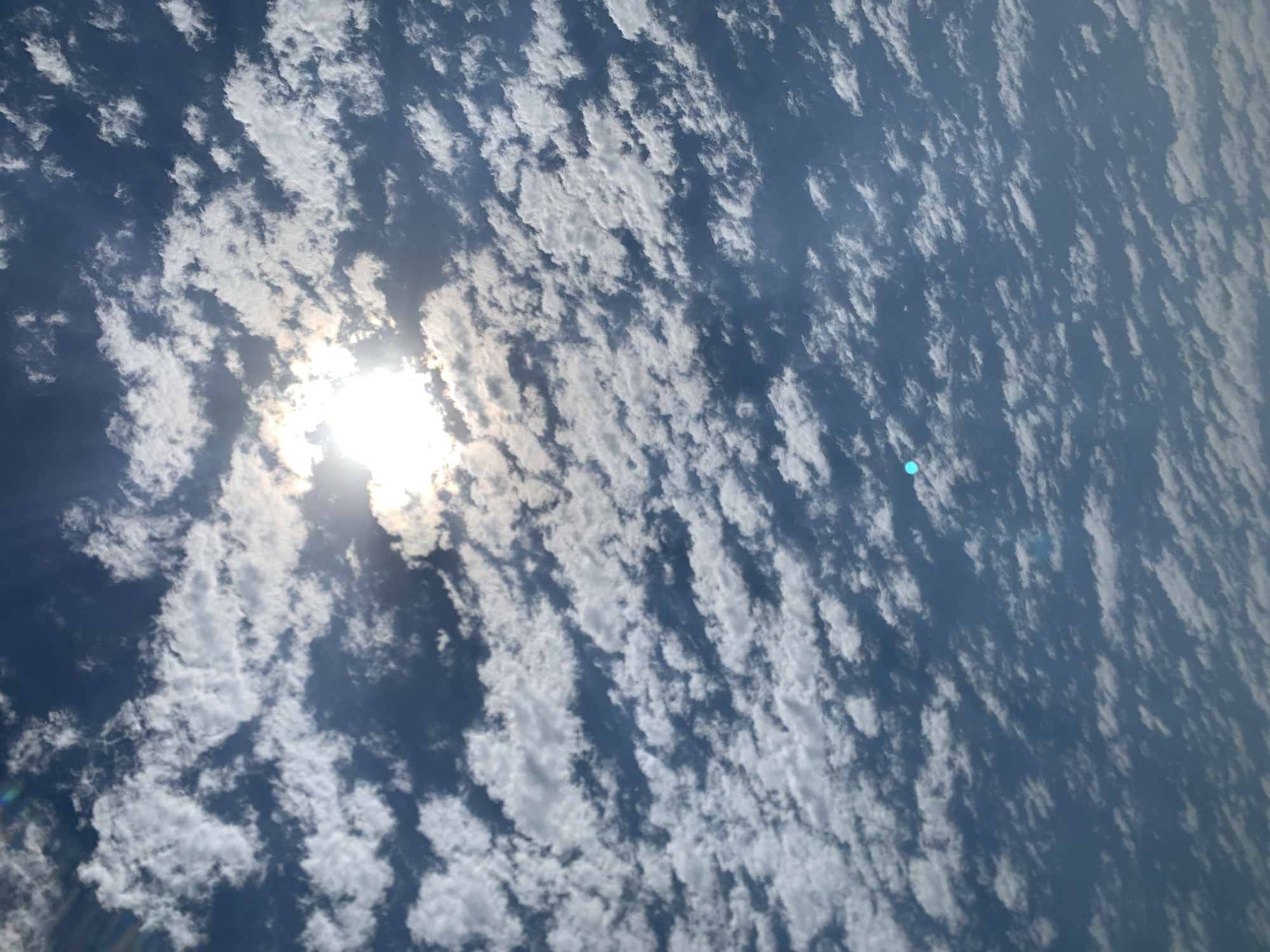 A low-angle shot captures a sky filled with dappled clouds against a blue backdrop. The sun is partially obscured by a cluster of clouds, creating a bright, diffused light. The clouds appear in various sizes and formations, with some appearing more wispy and others more defined. The sky is a clear, bright blue. There are no people or structures visible in the image.
