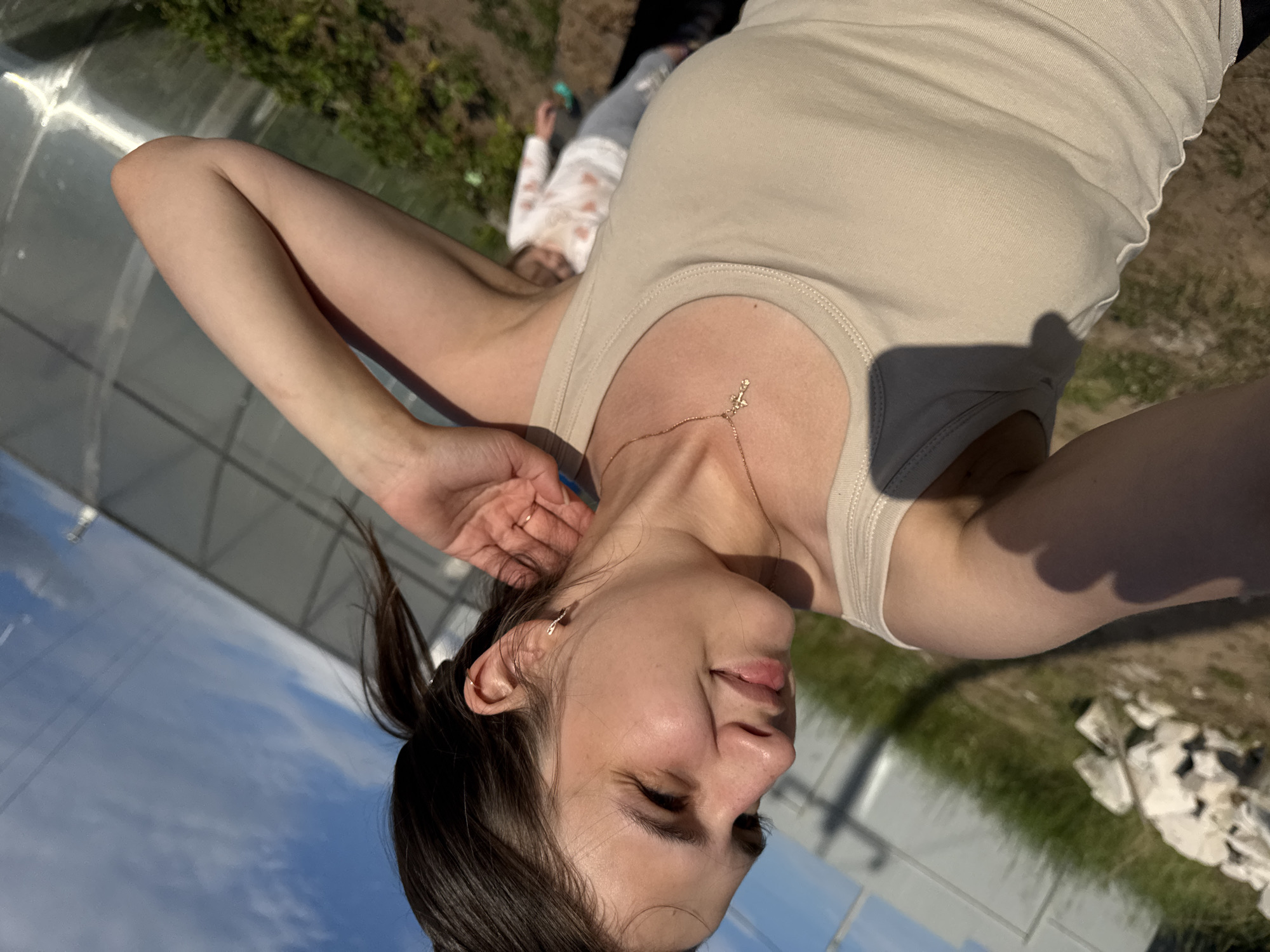 A young woman is captured in a selfie, her face turned towards the camera with a playful expression. She is wearing a beige tank top and a delicate gold necklace with a small cross pendant. Her left arm is raised, her hand resting on her head, and her hair is pulled back in a ponytail. In the background, a greenhouse with a transparent roof is visible, and beyond that, a patch of green grass and soil. The sky is blue with white clouds, suggesting it is daytime. Another person, likely a child, is lying on the grass in the background, wearing a white shirt with a pink floral pattern. The overall impression is a casual and relaxed moment captured outdoors, possibly in a garden or backyard. The lighting indicates it is likely a sunny day.