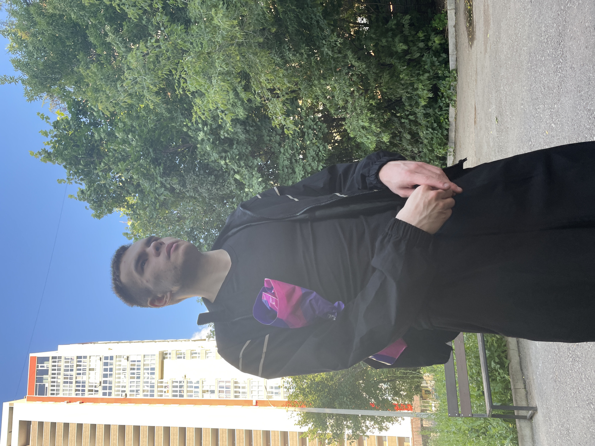 A young man stands outside in a courtyard on a sunny day, facing left and looking upwards. He is wearing a black t-shirt and a black zippered jacket. A colorful bag, featuring pink and purple stripes, is tucked into the front of his jacket. His hands are clasped together in front of him. In the background, a tall, white apartment building with orange accents dominates the left side of the frame, while lush green trees fill the right side. A wooden bench with a metal frame sits on the left, behind some tall grass. The ground is paved with asphalt. The sky is clear and blue.