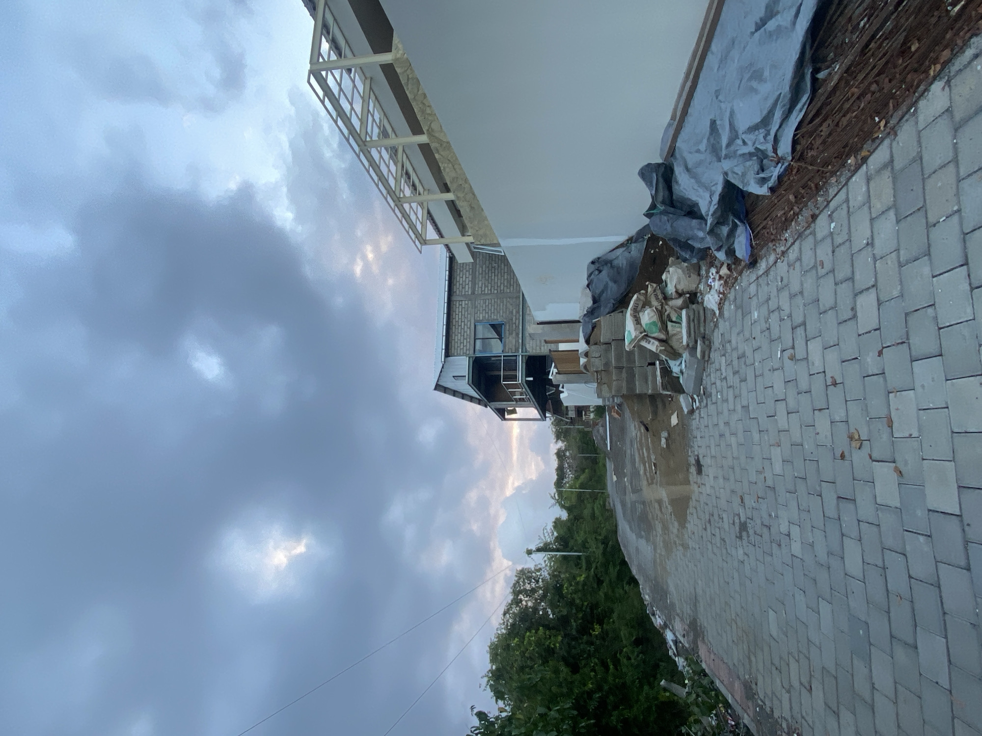 This is an outdoor, low-angle, full shot taken during the daytime in the city of Kuta Selatan, Indonesia. The scene depicts a narrow, paved road with a slight incline, leading uphill towards buildings under construction. The sky is overcast with heavy, dark clouds, but a patch of bright light indicates the sun is breaking through in the distance.
On the left side of the road, a dense cluster of green trees and foliage forms a natural boundary. A utility pole with visible wires is positioned near the trees. The road itself is paved with gray interlocking bricks, and in some areas, there are puddles of water and patches of exposed dirt, suggesting recent rain or ongoing construction. Scattered debris, including plastic bags, leaves, and small stones, are visible along the edge of the road.
On the right side of the road, a white stucco wall extends along the embankment. Above this wall, a multi-story building is under construction, featuring exposed concrete block walls, balconies with metal railings, and a skeletal roof structure made of metal beams. Several piles of construction materials are present, including bags of cement, stacks of bricks, and bundles of rebar covered with a dark blue tarp. The overall impression is a construction site in progress, possibly in a hilly or elevated area, under a dramatic, cloudy sky.