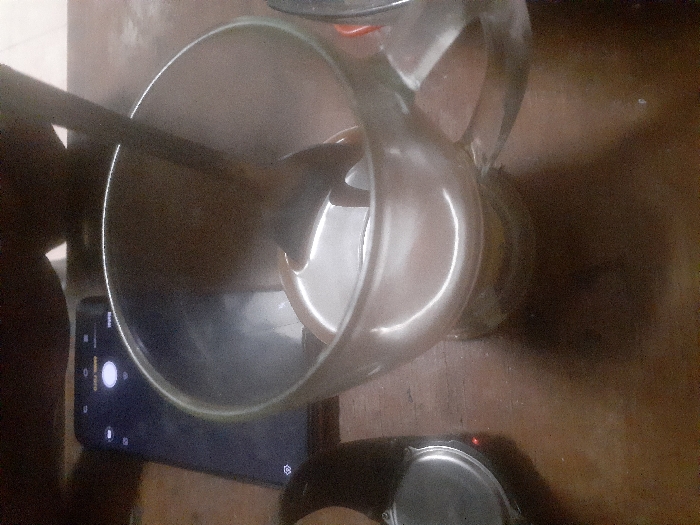 A top-down view depicts a clear glass mug containing a dark liquid and a spoon on a wooden surface in Cimenyan, Indonesia. To the left of the mug, a dark smartphone with its screen illuminated, displaying a camera application, rests horizontally. Further to the left, a portion of a smartwatch or similar wrist-worn device, featuring a metallic rear and a small red illuminated indicator, is partially visible. The wooden surface exhibits a polished or lacquered finish, reflecting light sources. The background is dark and out of focus, suggesting an indoor setting with ambient lighting. No individuals are visible.