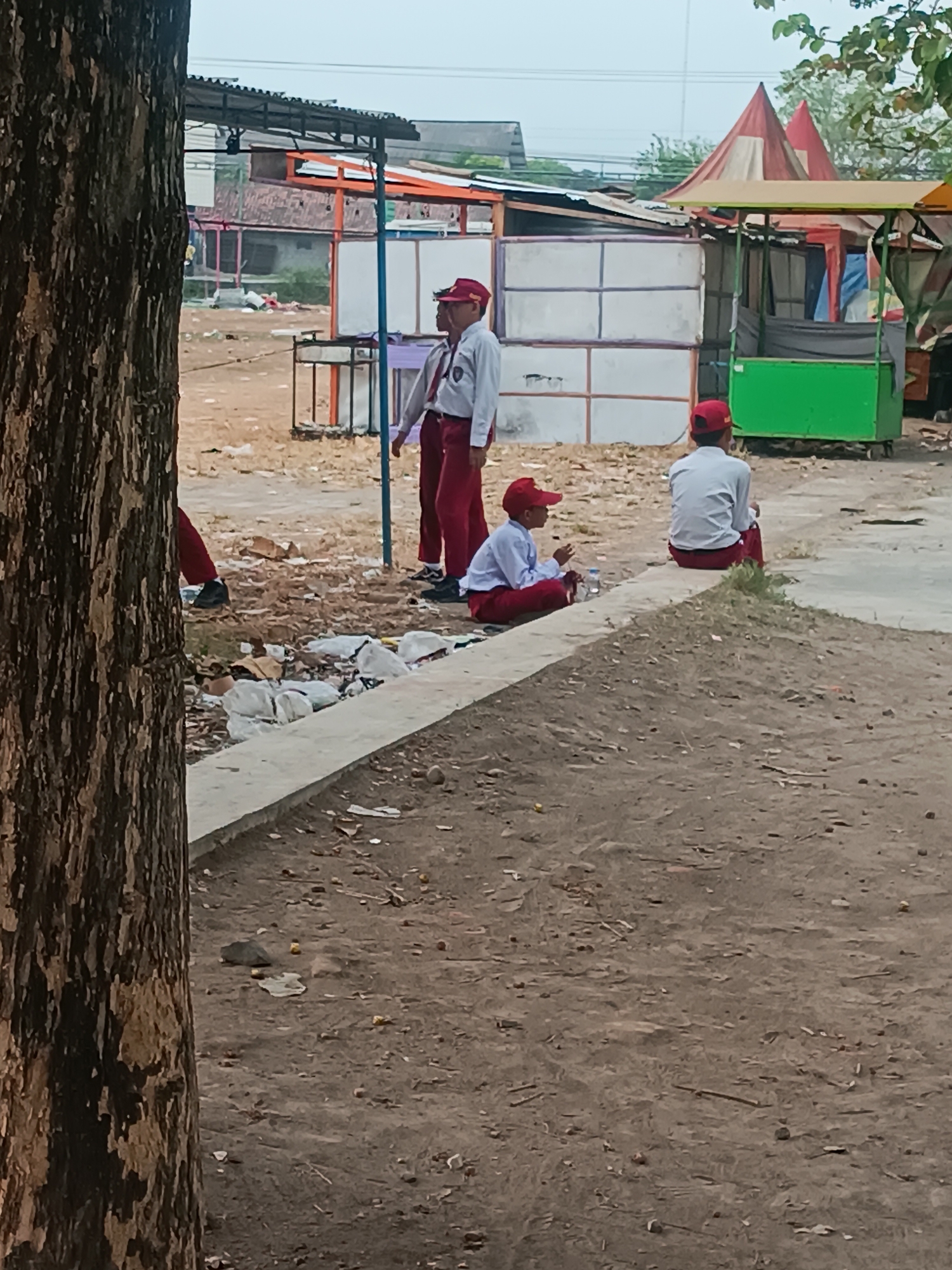 In the city of Pleret, Indonesia, an outdoor scene unfolds with several young students in uniform. In the foreground, a large tree trunk partially obscures the view on the left. Three students in red hats and white shirts with red ties are visible. One student stands facing left, with their back partially to the camera, wearing red pants and black shoes. Another student is sitting on a concrete ledge, facing left, holding something in their hands and wearing red pants and white shoes. A third student sits further back on the same ledge, facing away from the camera, wearing red pants and white shoes.
The environment appears to be a dusty, open area with several makeshift stalls or structures in the background. These structures have corrugated metal roofs and white paneling. A green cart-like structure is visible among the stalls. The ground is covered in dirt and debris, including scattered trash. The overall impression is one of a somewhat neglected outdoor space, possibly near a school or market. The sky is overcast.