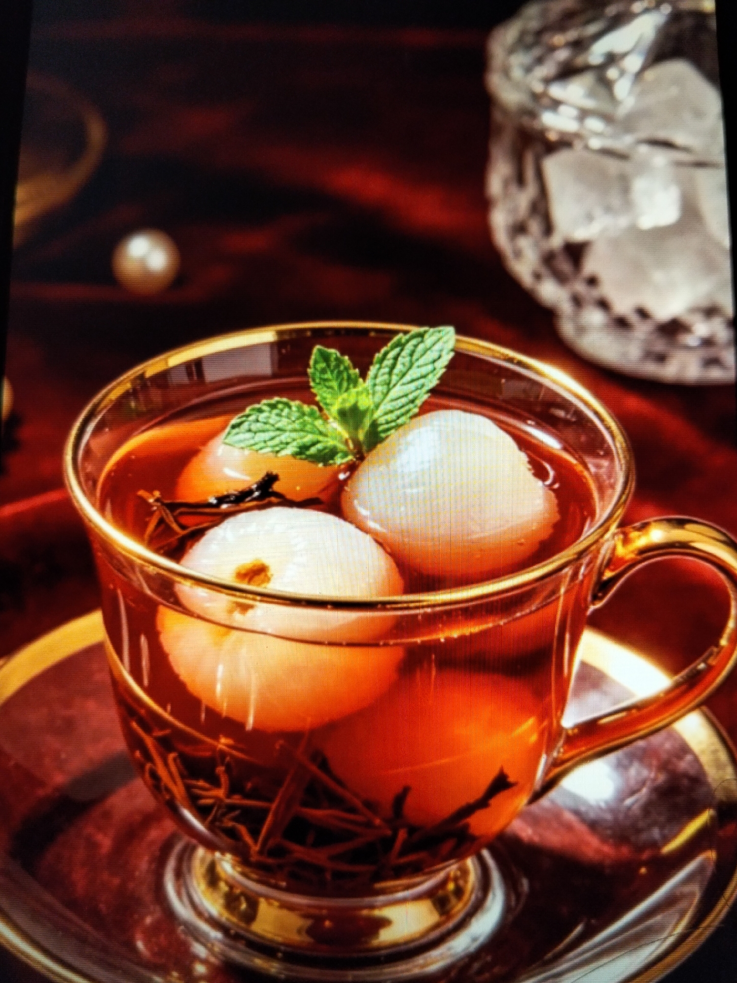 A close-up, dimly lit, high-angle shot displays a clear glass cup filled with amber-colored liquid and several peeled lychees. The cup has a gold rim and a gold handle, and it sits on a matching gold-rimmed saucer. A sprig of fresh mint rests on top of the lychees. In the background, out of focus, is a crystal decanter filled with ice cubes. A single pearl is visible on a deep red, draped fabric surface to the left. The overall aesthetic suggests a luxurious and refreshing beverage presentation. There are no people present in the image. The scene does not provide any information about a location in Payagaboh, Indonesia.