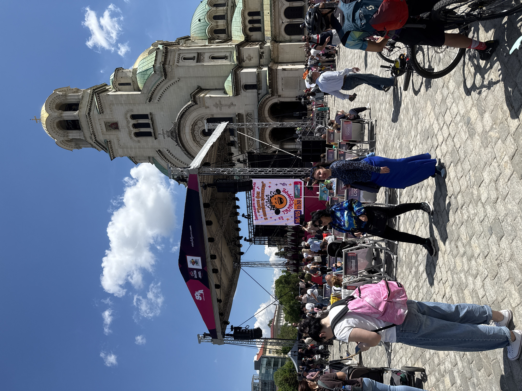 Here is a detailed description of the image:
**Setting:** The image appears to be taken in a large public square in Sofia, Bulgaria. The Alexander Nevsky Cathedral, with its distinctive golden domes, is prominently visible in the background, indicating the central location.
**Subjects and People:**
* Multiple people of various ages and appearances are present. Some are standing, some are seated in chairs, and others appear to be walking.
* One woman in a blue dress stands prominently in the foreground.
* A person with a pink backpack looks down while checking their phone.
* Some people are riding bicycles in the background.
**Objects:**
* A large stage is set up in the square, with a backdrop featuring text in Bulgarian, musical notes, and a cartoonish graphic.
* Chairs are arranged in rows facing the stage, suggesting an audience area.
* Lenovo brand chairs.
* A banner with the inscription "visitBulgaria.com" can be seen on the left side of the image.
**Actions and Interactions:**
* People are gathered in the square, likely for an event taking place on the stage.
* The woman in blue looks towards the camera.
* There seems to be some type of festival.
**Environment:**
* The weather appears to be sunny, with blue skies and some white clouds.
* The ground is paved with stone blocks.
* Trees and buildings are visible in the surrounding area, contributing to the urban setting.