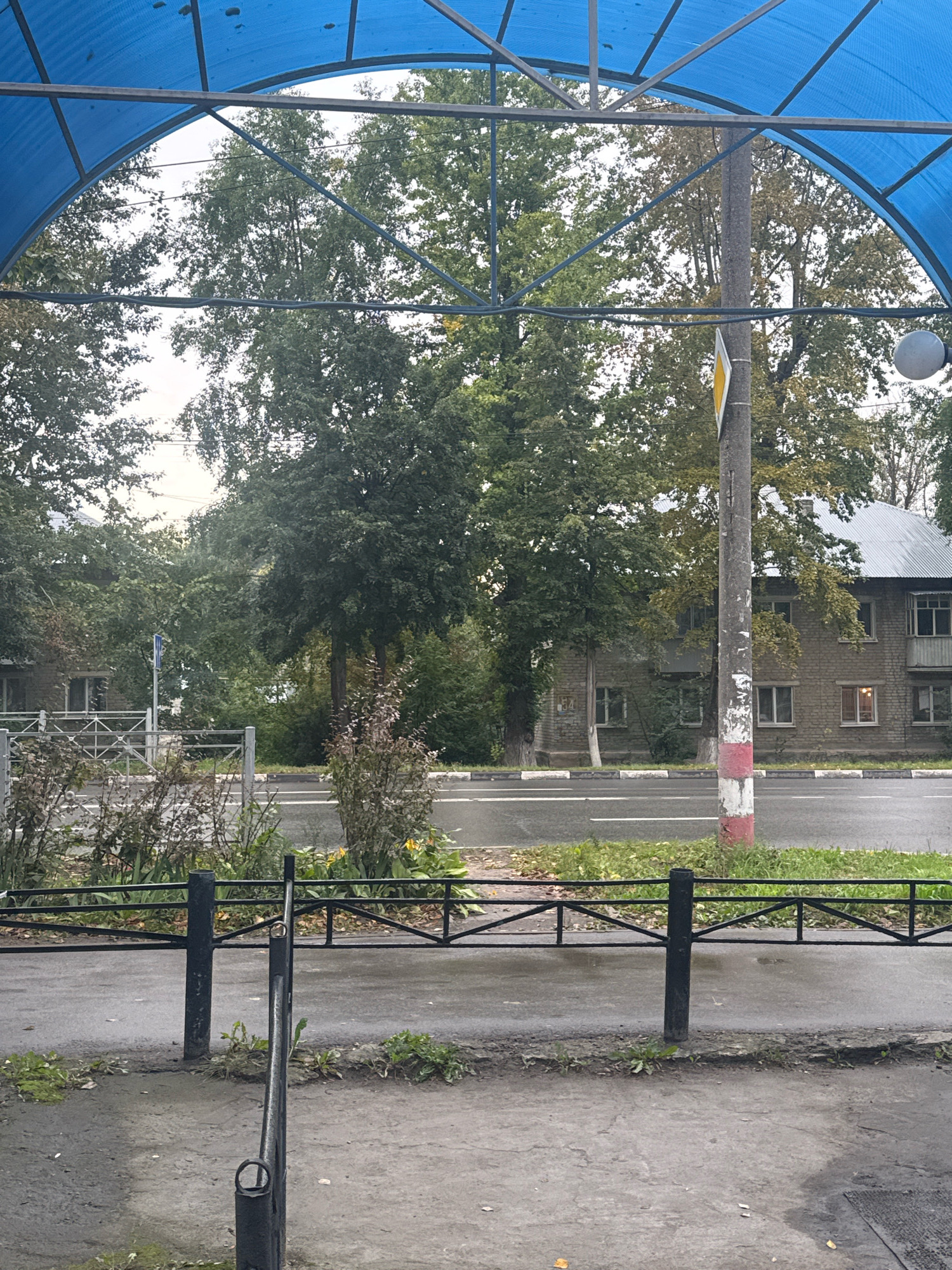 The image shows a street scene viewed from under a blue awning. The awning is supported by metal beams. Through the awning, a street is visible with trees lining the opposite side. A multi-story brick building with a metal roof is partially obscured by the trees. A lamppost with red and white markings stands near the street. A black metal fence runs along the side of the road. The pavement appears wet, suggesting it may have rained. The scene is likely a residential or commercial area within the city of Ulyanovsk, Russia, given the architectural style and general environment. There are no people or animals visible in the image.