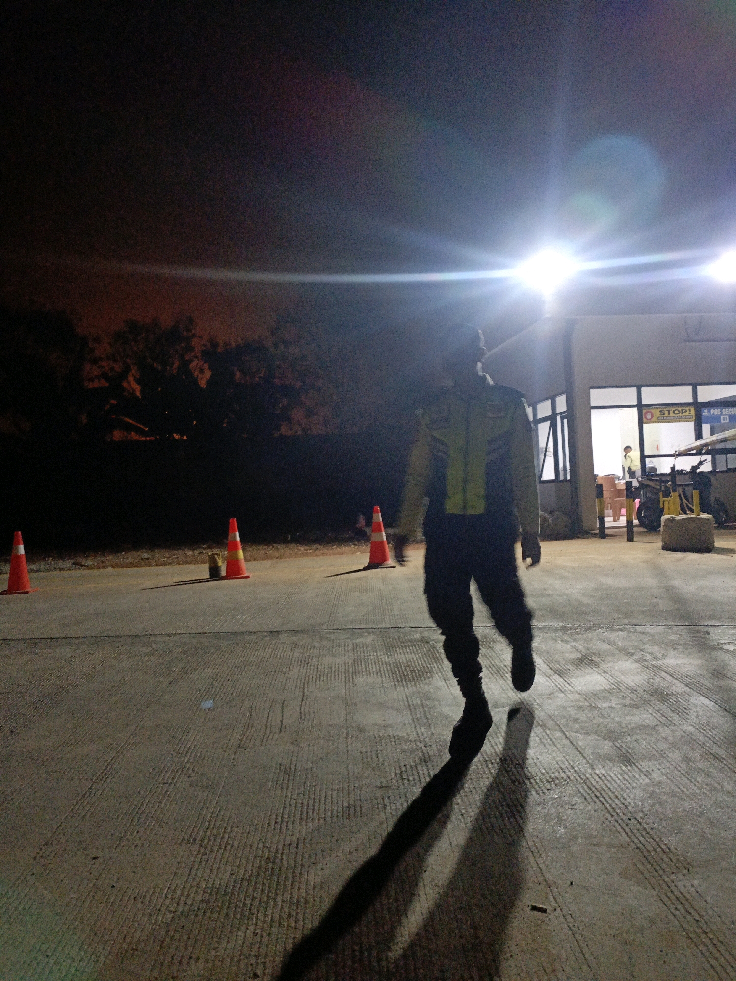 A security guard, silhouetted and facing away from the camera, walks across a concrete surface at night. The guard is wearing a high-visibility jacket with reflective stripes over dark pants. Bright lights illuminate the scene from the right, casting a long shadow of the guard on the ground. In the background, a building with windows and a sign reading "STOP!" is visible. Orange traffic cones are placed on the ground at intervals. Trees are dimly visible in the dark background. The overall scene suggests an outdoor industrial or facility area. The location is identified as Jenu, Indonesia.