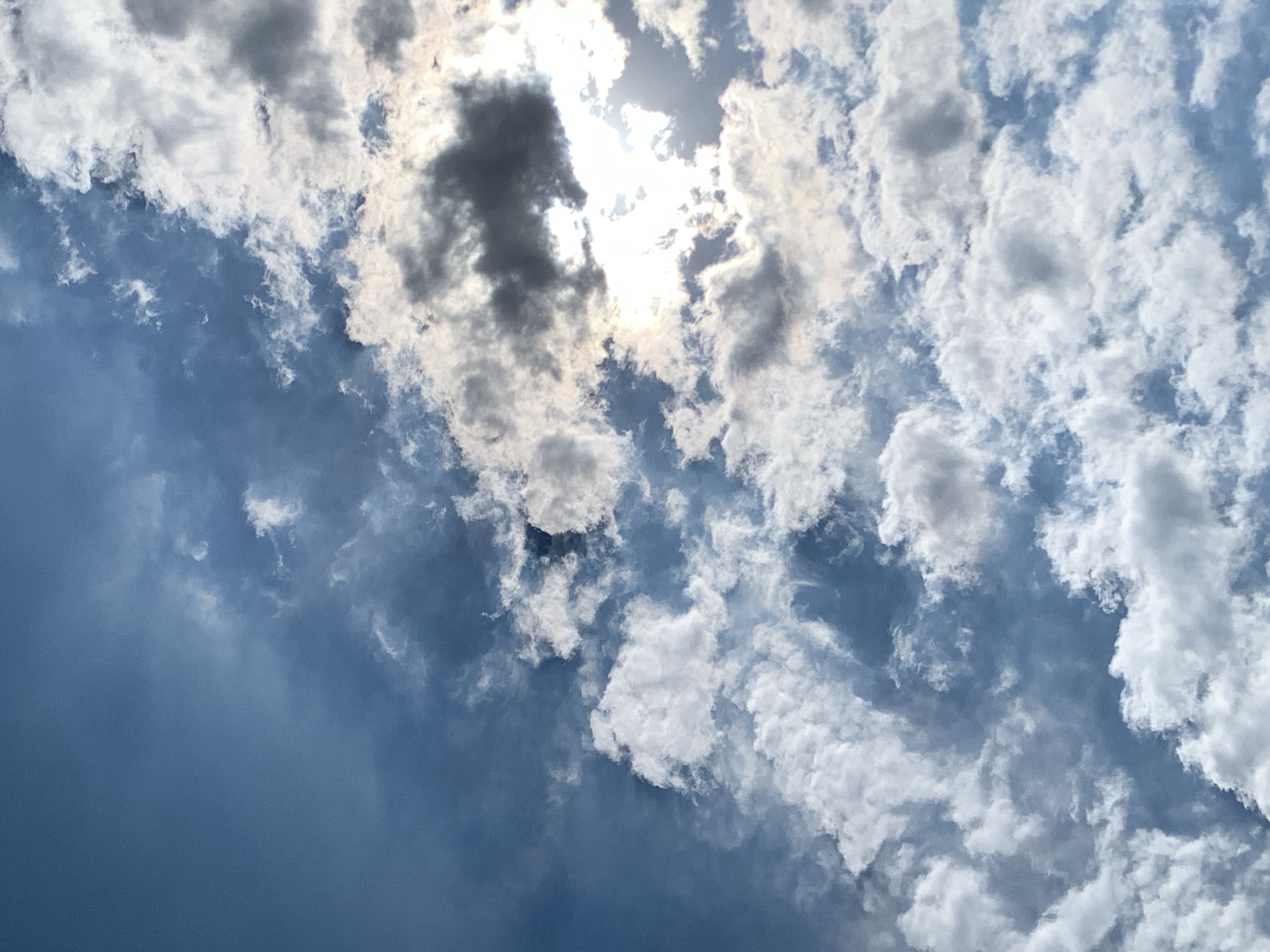 The image shows a sky filled with a variety of clouds, ranging from bright white and fluffy cumulus clouds to darker, more ominous storm clouds. The sun is visible, peeking through a gap in the clouds, casting a bright light and creating highlights on the cloud edges. The sky is a deep blue, with some areas appearing slightly hazy. The perspective is looking upwards, suggesting the photo was taken from ground level. There are no people, structures, or any other elements of a city visible in the frame. The image solely focuses on the atmospheric conditions.