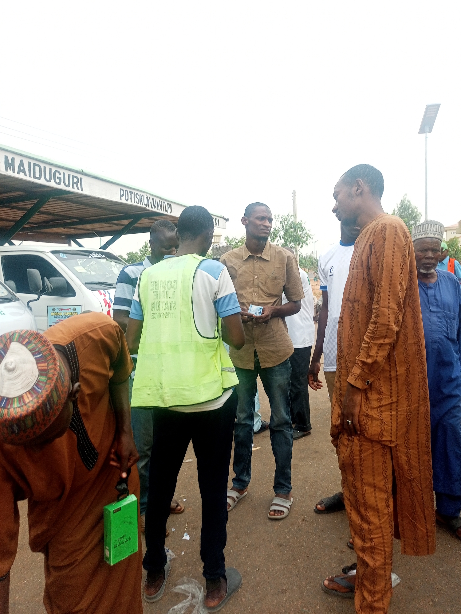 A group of men are standing on an asphalt surface under an overhang labeled "MAIDUGURI" and "POTISKUM-DAMATURU," in an outdoor setting. One man is wearing a high-visibility vest labeled "COMBO EXPRESS LINE STATIONERS." Another man is holding cash in his hand. At least one other individual is holding a green rectangular box. Some men are wearing traditional clothing. A white van is visible in the background. The scene is in Gombe, Nigeria.