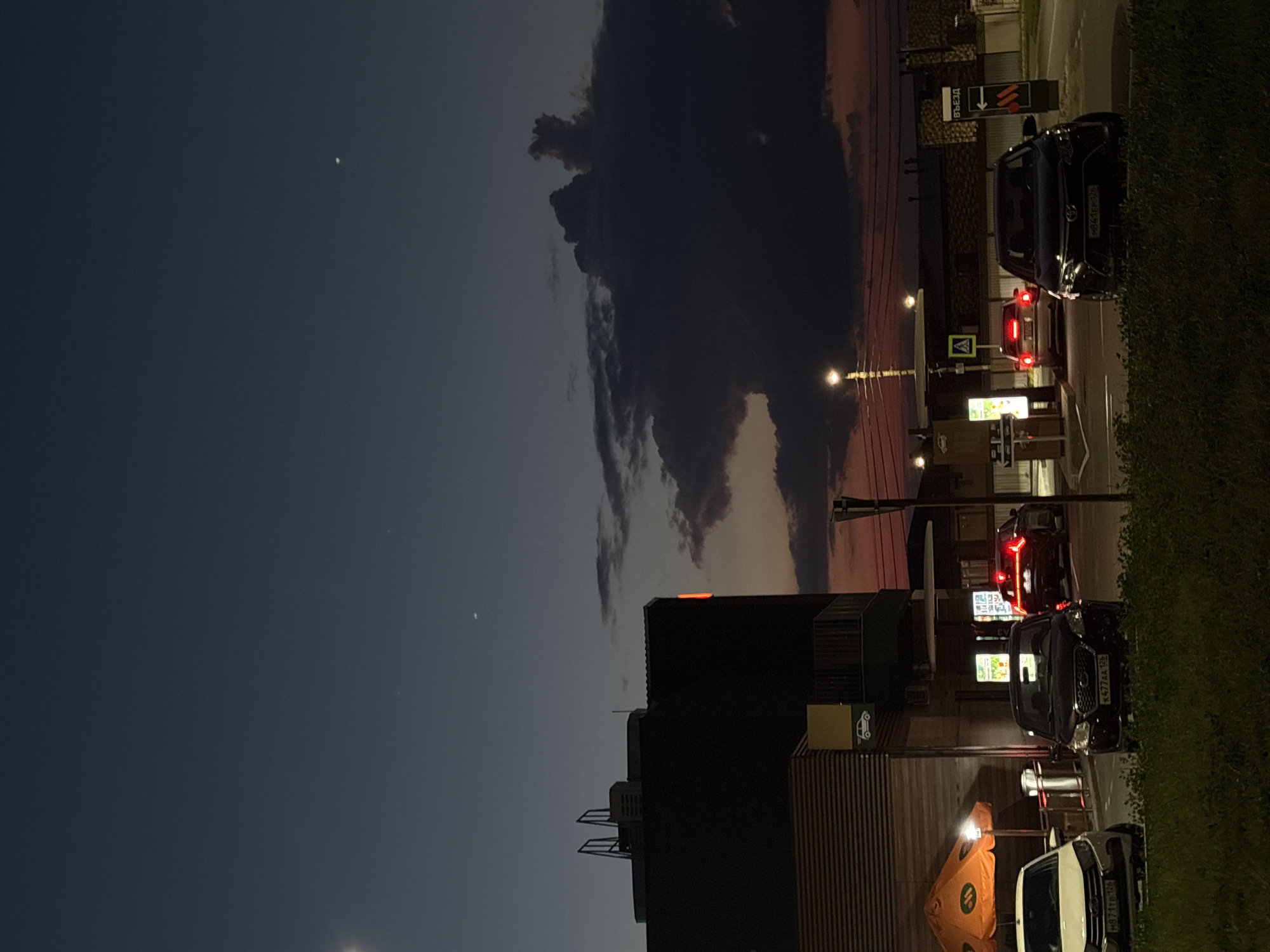 The media file shows a street scene at dusk in Mikhaylovsk, Russia. The sky is dark blue with some reddish hues near the horizon, indicating sunset. Dark clouds are present, and there are a few bright points of light, possibly stars or distant lights.
Several cars are visible on the street. One dark-colored SUV is parked on the right side of the image, facing away from the viewer. Its taillights are illuminated. Further down the street, another dark car is seen, also with its taillights on, suggesting it is either stopped or moving slowly. In the foreground, there's a white car with its back to the viewer. Adjacent to it is an orange awning.
Buildings line the right side of the street. One building appears to be a commercial establishment with signage. There are streetlights and what look like traffic signals. A sign with Cyrillic text "ВЪЕЗД" (meaning "entrance") is visible on a pole. Another sign shows an arrow pointing down and some orange circular symbols. A yellow sign with a car icon is also visible.
The scene appears to be a quiet street, possibly near a commercial area. The time of day is clearly dusk or early evening, given the sky conditions and the illuminated car lights. The lighting is dim, with artificial lights from the street and buildings adding to the scene. No people are clearly visible.