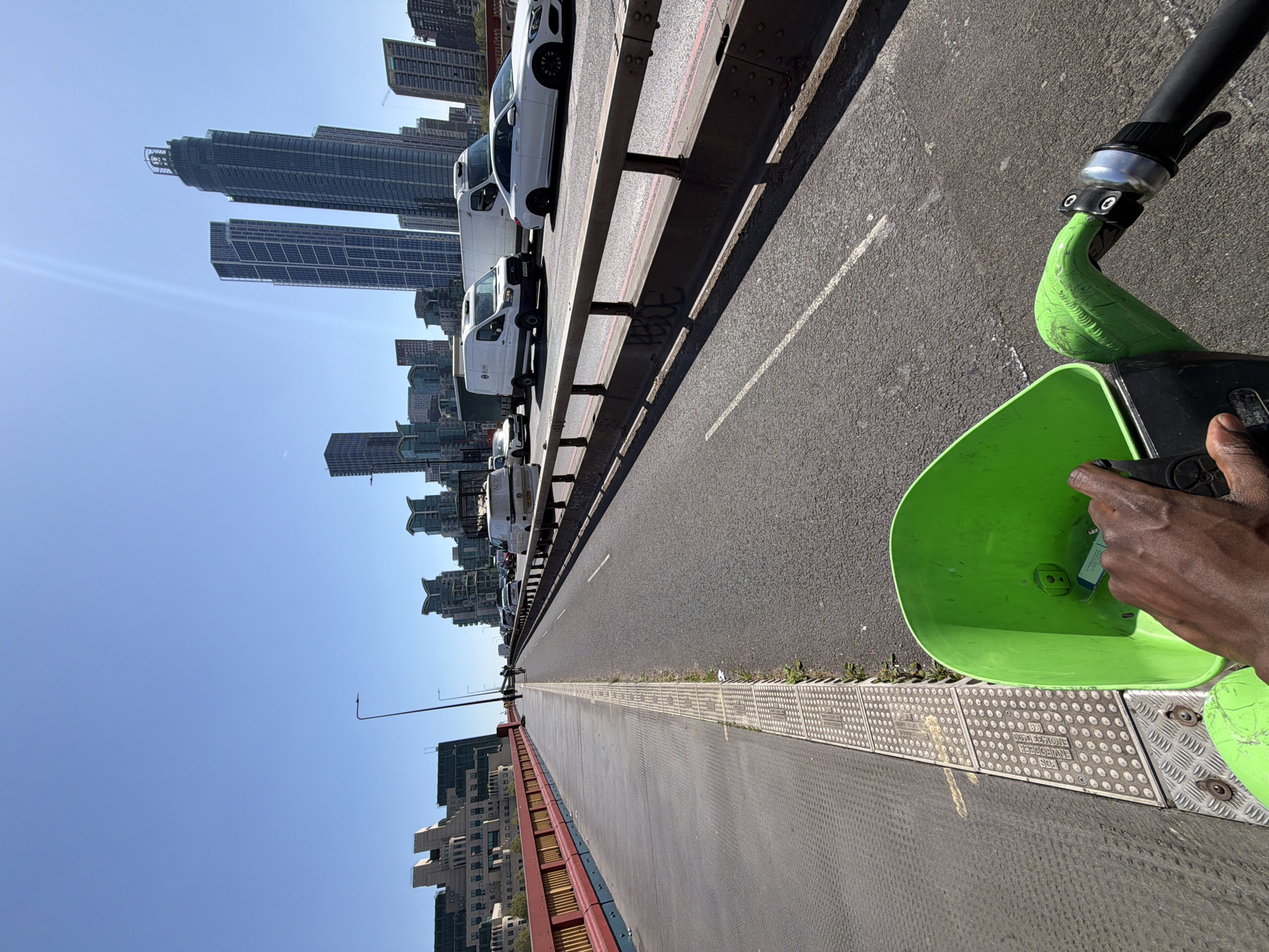 This image, taken on a clear, sunny day in what appears to be a major urban center like London, United Kingdom, shows a first-person perspective from an electric scooter. A dark-skinned hand is visible in the foreground, holding an object inside the bright green front basket or compartment of the scooter. Part of the scooter's black handlebar and green body are also visible.
To the right of the scooter, a pedestrian or cycle path features textured tactile paving panels, some of which clearly display the embossed text "ENVIRODECK" and "SPS". To the left, a sturdy concrete barrier separates this path from a roadway. A line of white cars and vans is visible on the road, suggesting traffic or parked vehicles. The text "UK PADS" is partially visible on the side of one white van. Graffiti spelling "MORSE" is spray-painted on the concrete barrier.
In the background, under a vast, bright blue sky with a visible sun flare, a dense collection of modern skyscrapers forms a prominent city skyline. The clear skies and strong shadows indicate bright midday conditions. The scene suggests a bustling urban environment, likely on a bridge or elevated road, with the person on the scooter moving through or observing the city.