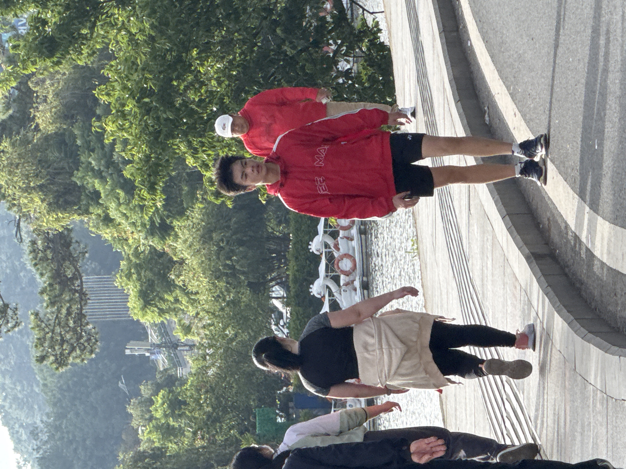 This image depicts a daytime outdoor scene on a paved pathway adjacent to a body of water, likely Xuan Huong Lake in Da Lat, Vietnam, given the characteristic swan pedal boats and the mountainous, tree-covered background.
On the paved pathway, five individuals are visible, walking from left to right.
1. **Far left (partially obscured):** The arm and side of a person wearing dark clothing are visible, suggesting they are walking alongside another woman.
2. **Second from left:** A woman with her back mostly to the viewer, wearing a light-colored top over dark trousers. Her head is turned slightly to the right.
3. **Center-left:** A woman with dark hair tied in a ponytail, facing away from the viewer. She is wearing a black short-sleeved top with grey raglan sleeves and black leggings. A light beige/cream hooded jacket is tied around her waist. She wears light-colored sneakers with a visible pink detail on one shoe.
4. **Center-right:** A young man with dark, spiky hair, walking towards the viewer's left. He wears a red hooded windbreaker-style jacket with white stripes on the sleeves and text on the chest that appears to read "EE MA". He has black athletic shorts (possibly Yonex brand) and dark sneakers with white socks.
5. **Far right:** A man walking behind the young man, facing away from the viewer. He wears a red hooded sweatshirt and light-colored (beige/khaki) pants. He has a white baseball cap with a green crocodile logo (Lacoste) on it and carries a white bottle or container in his right hand. He is wearing white sneakers.
To the left of the pathway is a body of water where several white swan-shaped pedal boats with orange lifebuoys are moored. The pathway itself is light grey concrete with distinct horizontal tactile paving lines and a darker curb separating it from an asphalt road on the right, which is mostly out of frame.
The background features dense green trees and foliage, rising into a mountainous or hilly landscape, characteristic of Da Lat. In the distant background, amidst the trees, a tall, multi-story building with vertical architectural lines is visible, along with what appears to be a telecommunications tower. A blue sign with white text, partially reading "RIV", is discernible above the trees in the upper right background. The scene is illuminated by strong sunlight, casting shadows on the ground, suggesting either morning or late afternoon.