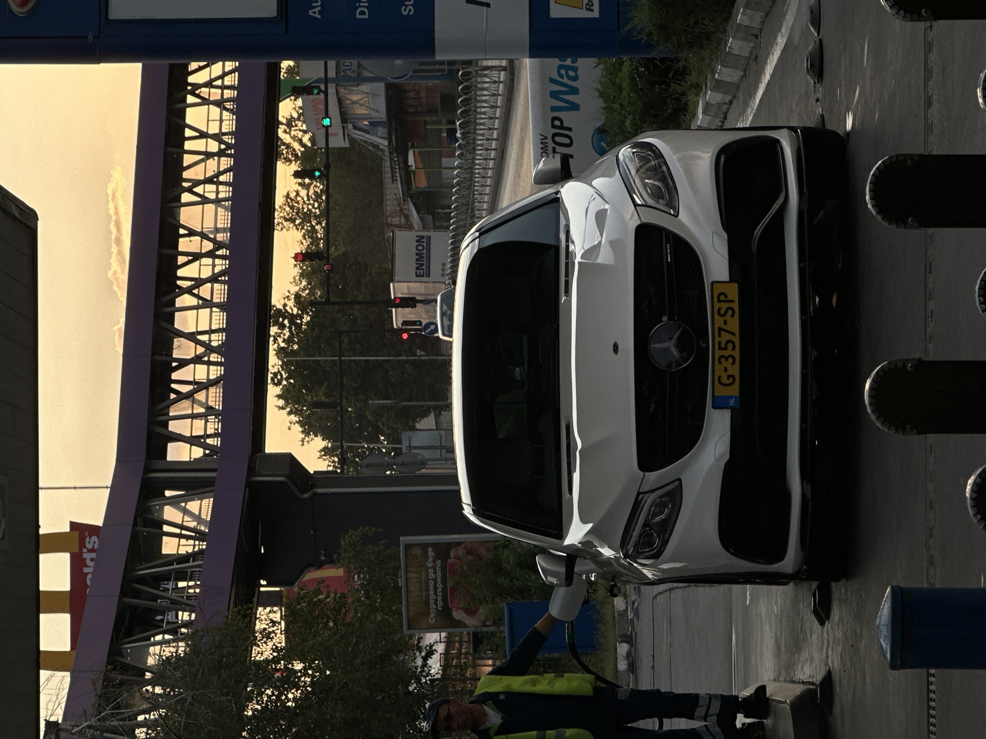 The image captures a white Mercedes-Benz GLE at a gas station in Veliko Tarnovo, Bulgaria, under a dusky sky. A gas station attendant, wearing a yellow vest and blue uniform, is visible on the left, holding a gas nozzle near the car. The car's license plate reads "G-357-SP" with "NL" indicating its Dutch registration. In the background, a McDonald's sign is prominent on a purple pedestrian overpass. Traffic lights and other structures associated with a city environment are also present, with trees and foliage indicating an outdoor setting. The overall scene suggests a typical urban day at a refueling station.
