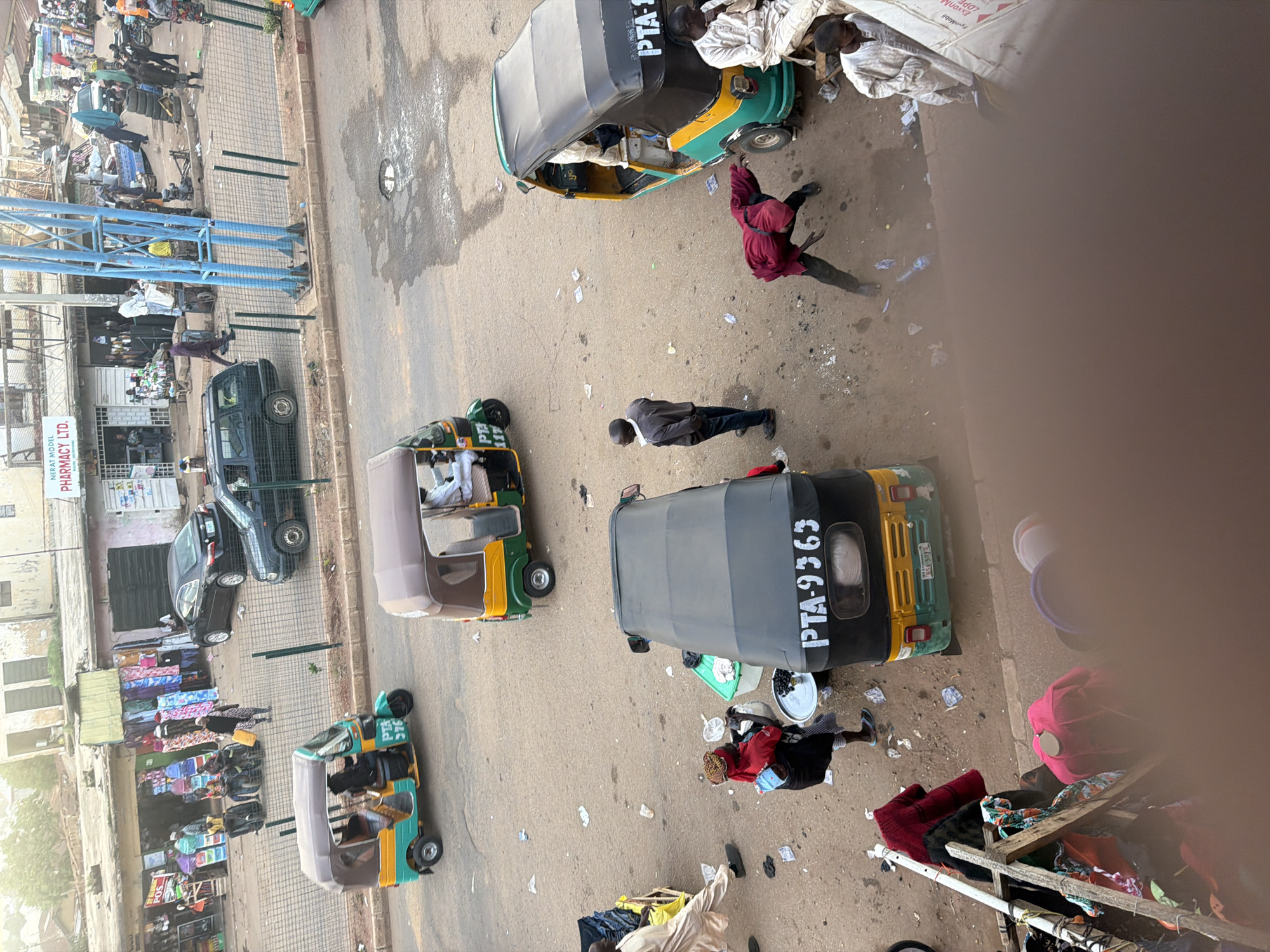 This media file captures a bustling street scene in Bukuru, Nigeria. The image is dominated by several auto-rickshaws, commonly known as "Keke Napep," suggesting a lively transportation hub. One Keke Napep is prominently displayed with the text "PTA-9363" visible on its side, indicating a vehicle registration or identification. Another Keke Napep has "PTA 117" and "RSA SOYB" on its rear. In the background, there is a building with a sign that reads "NERAT MODEL PHARMACY LTD."
People are scattered throughout the scene. Some are standing near or in the Keke Napeps, possibly passengers or drivers. Others appear to be pedestrians on the sidewalk or interacting with street vendors. One person in a red garment is captured in motion, adding a dynamic element to the composition. There are also piles of goods, likely merchandise being sold on the street, with textiles and bags visible. The ground is paved, and there are some scattered debris, typical of a busy urban environment. The lighting suggests it might be daytime, perhaps late morning or early afternoon, with the sun casting shadows. The overall impression is one of everyday life and commerce unfolding in a Nigerian town.