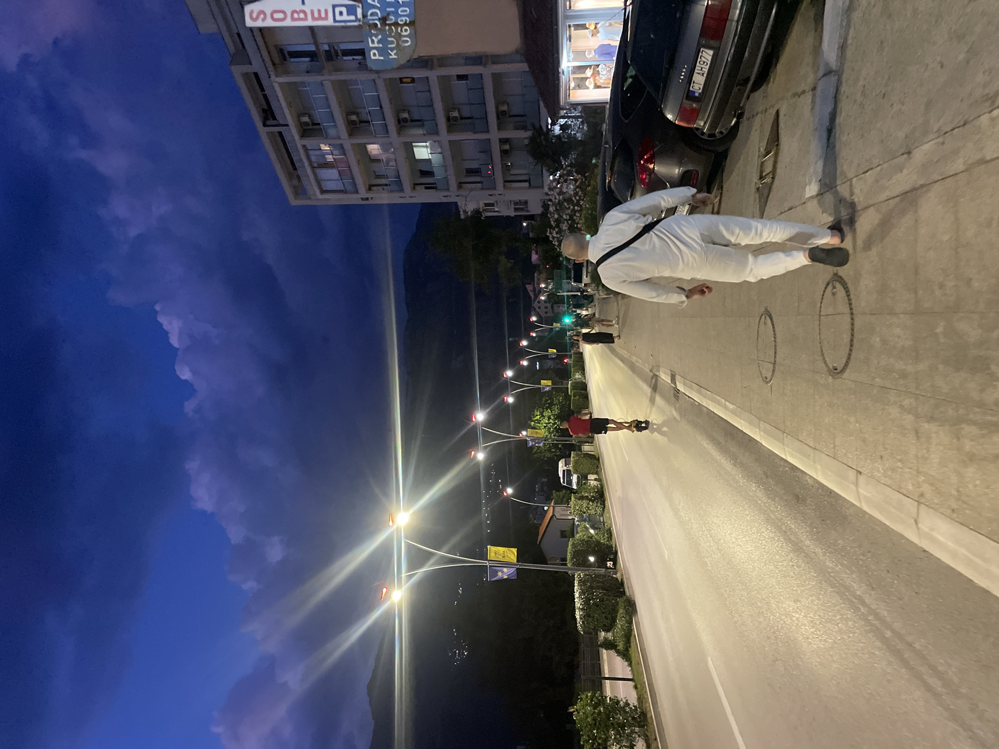 This is a medium-shot, eye-level, outdoor, wide-angle photograph taken at dusk on a street in Budva, Montenegro. A man in a white linen outfit walks away from the camera on the right side of the image, heading towards the right. He has a black bag slung over his left shoulder. To the left of the sidewalk, several cars are parked. A dark sedan with license plate CT AH977 is visible on the far right. Further left, a light gray sedan and a darker colored SUV are parked. The sidewalk is paved with light-colored square tiles, and several circular manhole covers are visible.
Across the street, a man on a yellow electric scooter is riding away from the camera towards the left. He is wearing a red t-shirt and dark shorts. The street is paved with asphalt and appears wet, reflecting the streetlights. Several lampposts with bright lights are evenly spaced along the left side of the road, with two flags hanging from one of them. Beyond the road, there is a row of manicured green bushes. In the background, mountains are visible under a dark blue sky with dramatic clouds and streaks of light.
To the right of the parked cars, a multi-story building with balconies and air conditioning units is visible. A sign on the building, partially obscured, reads "SOBE P" and below that "PRODAJA KUĆA 06901". Adjacent to the building, a storefront with mannequins displaying clothing is visible. The overall scene suggests a residential or commercial street at the end of the day.