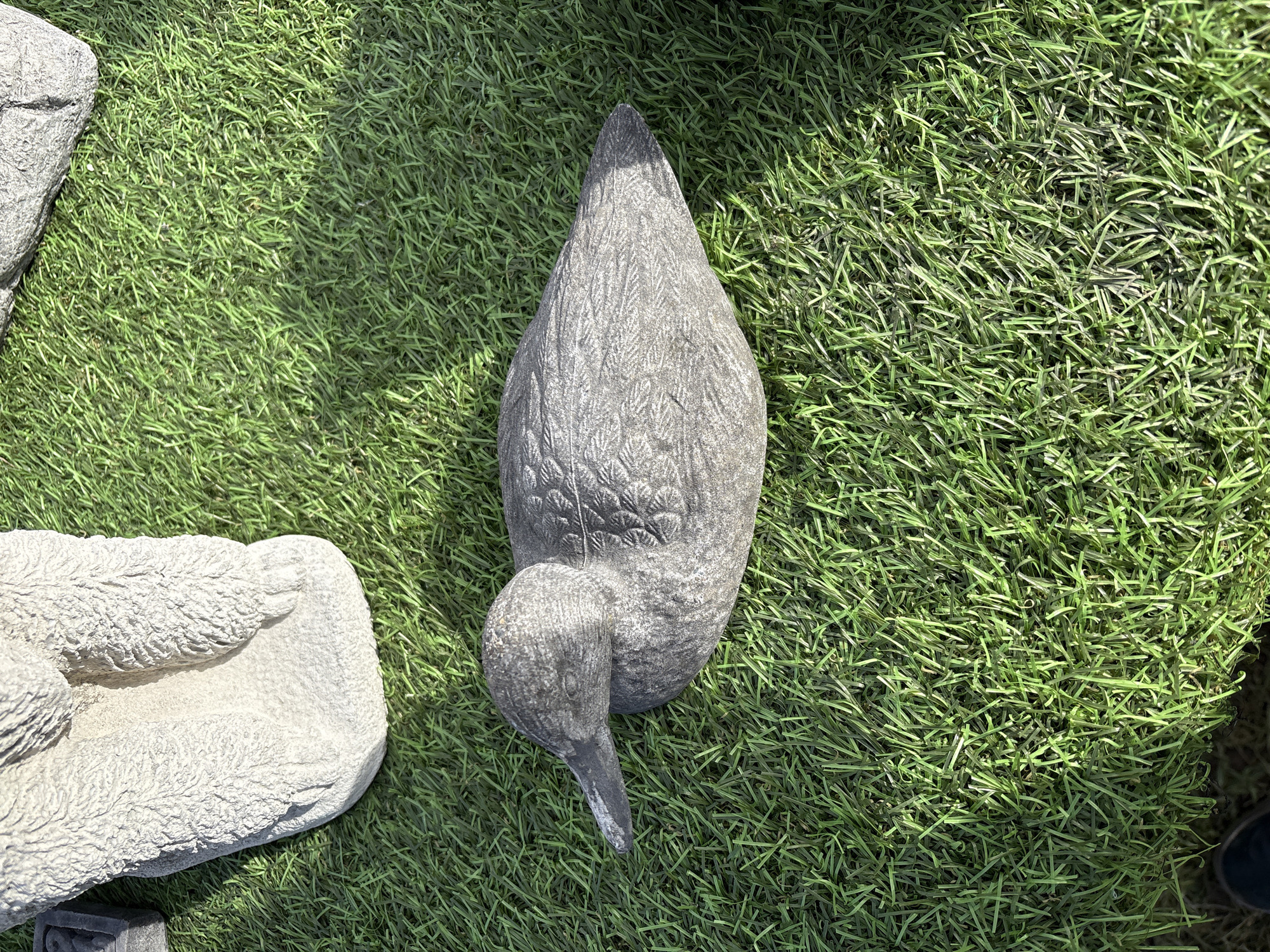 The image shows a gray concrete bird sculpture lying on green artificial turf. The sculpture depicts a bird with its head turned down and to the left. Another concrete sculpture of an animal rests partially visible on the left edge of the frame. A small portion of a dark shoe is visible in the bottom right corner. The scene is outdoors and well-lit. Based on information available, this image was taken in Chirk, United Kingdom.