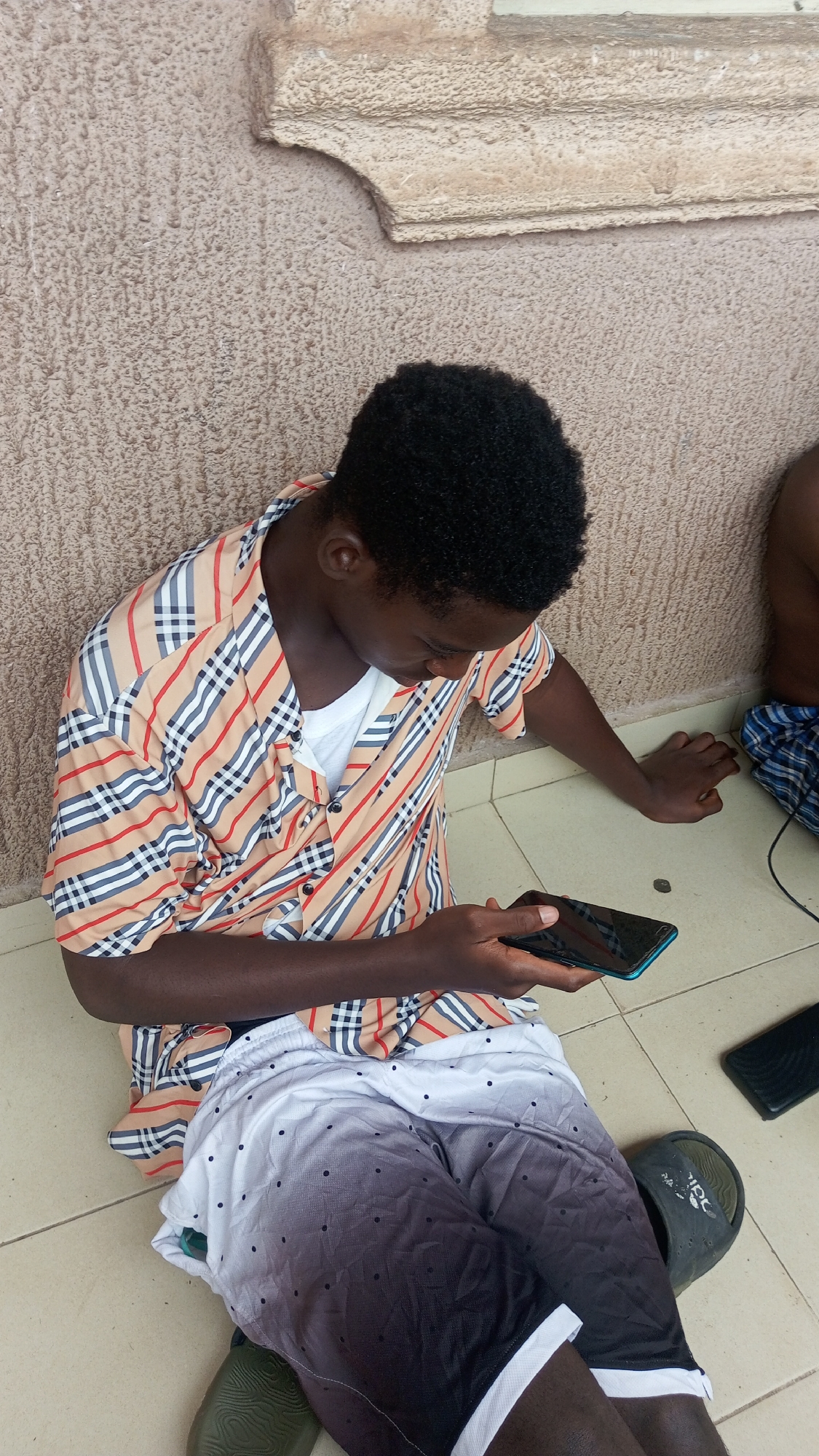 A person is seated on a tiled floor, engrossed in a black smartphone with a blue/teal edge. They have dark skin and short, dark, curly hair, and are looking down at the phone screen. They are wearing a short-sleeved, collared shirt with a plaid pattern featuring tan, red, black, and white stripes, over a white t-shirt. Their shorts are white at the top, gradient to dark grey/black at the bottom, and have small black polka dots. Dark green slide sandals are visible on their feet.
The setting appears to be indoors or in a covered outdoor area, with light-colored square floor tiles and a textured, light brown/beige wall. A decorative architectural molding is visible on the wall above the person's head. On the floor to the right, another dark rectangular object, possibly a phone or power bank, is visible, along with a small, dark, circular object. A partial view of another person, showing their leg covered by a blue and white patterned cloth, is visible on the far right of the frame, suggesting they are also seated nearby. The lighting is bright and even, indicating daytime. The person appears focused and calm while using their device. No legible text is visible.