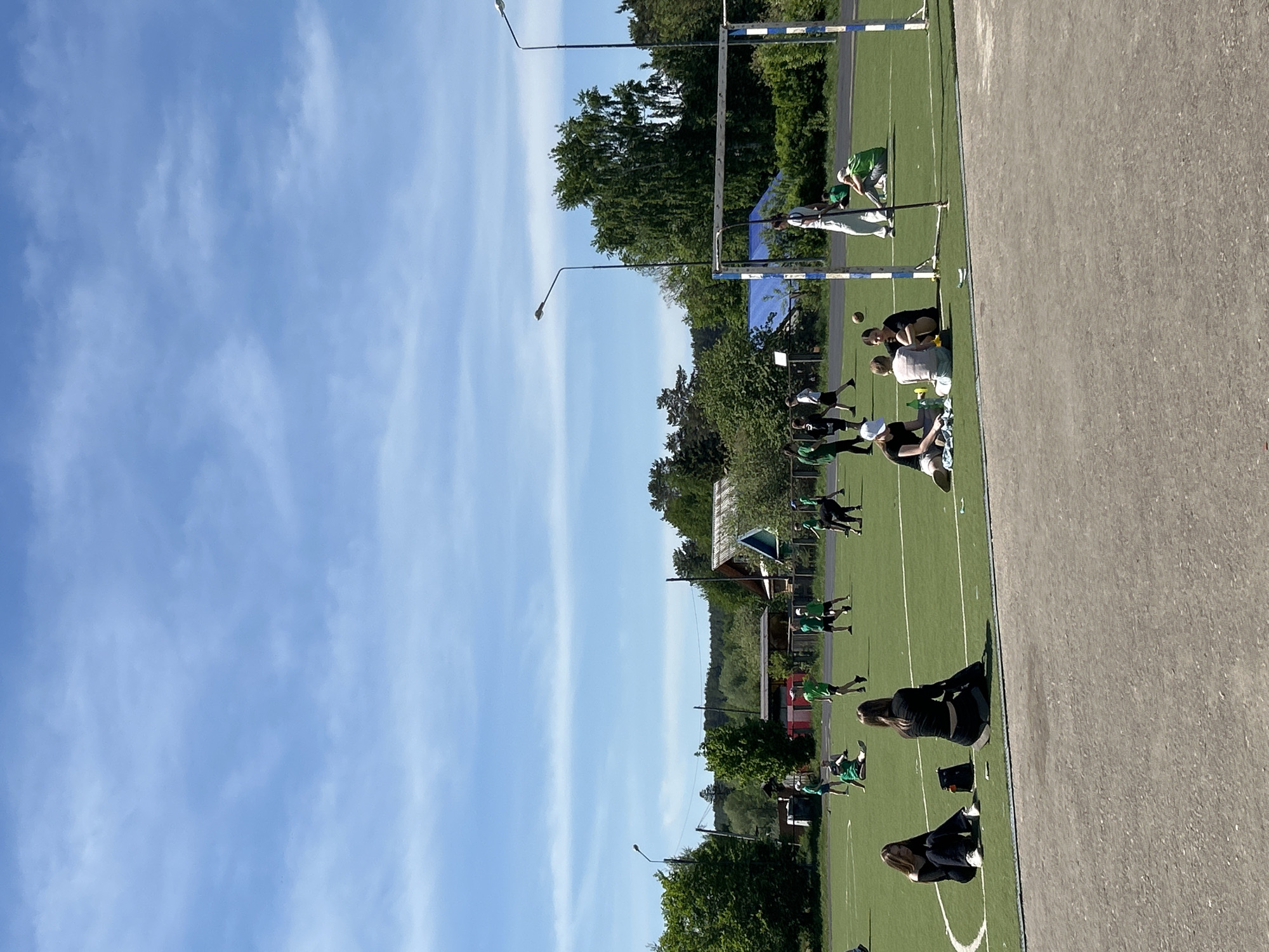 The image shows a scene at the "Snezhinka" stadium in Chaikovsky, Russia. A group of people are on a green field; some appear to be athletes stretching or warming up in athletic wear, while others are sitting or resting. A goal post is visible. The sky is blue with some clouds. A building is visible in the background.