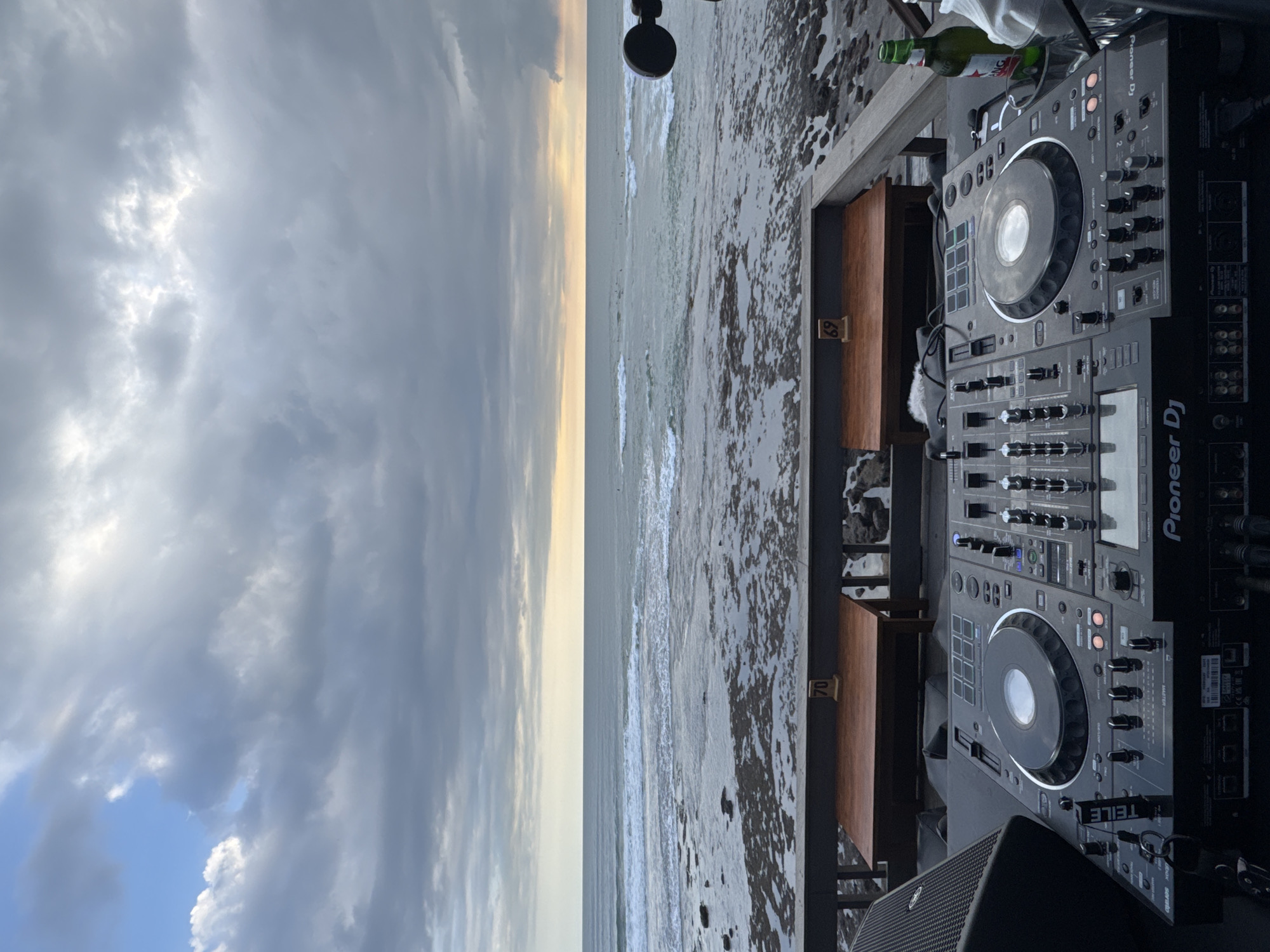 From a high angle, a Pioneer DJ set is situated outdoors, overlooking the ocean. A speaker is placed to the lower left, and a bottle of wine is near the top, to the right of the mixing equipment. To the left, the beach stretches away, fading into the horizon. The sky is cloudy. The location is North Kuta, Indonesia.