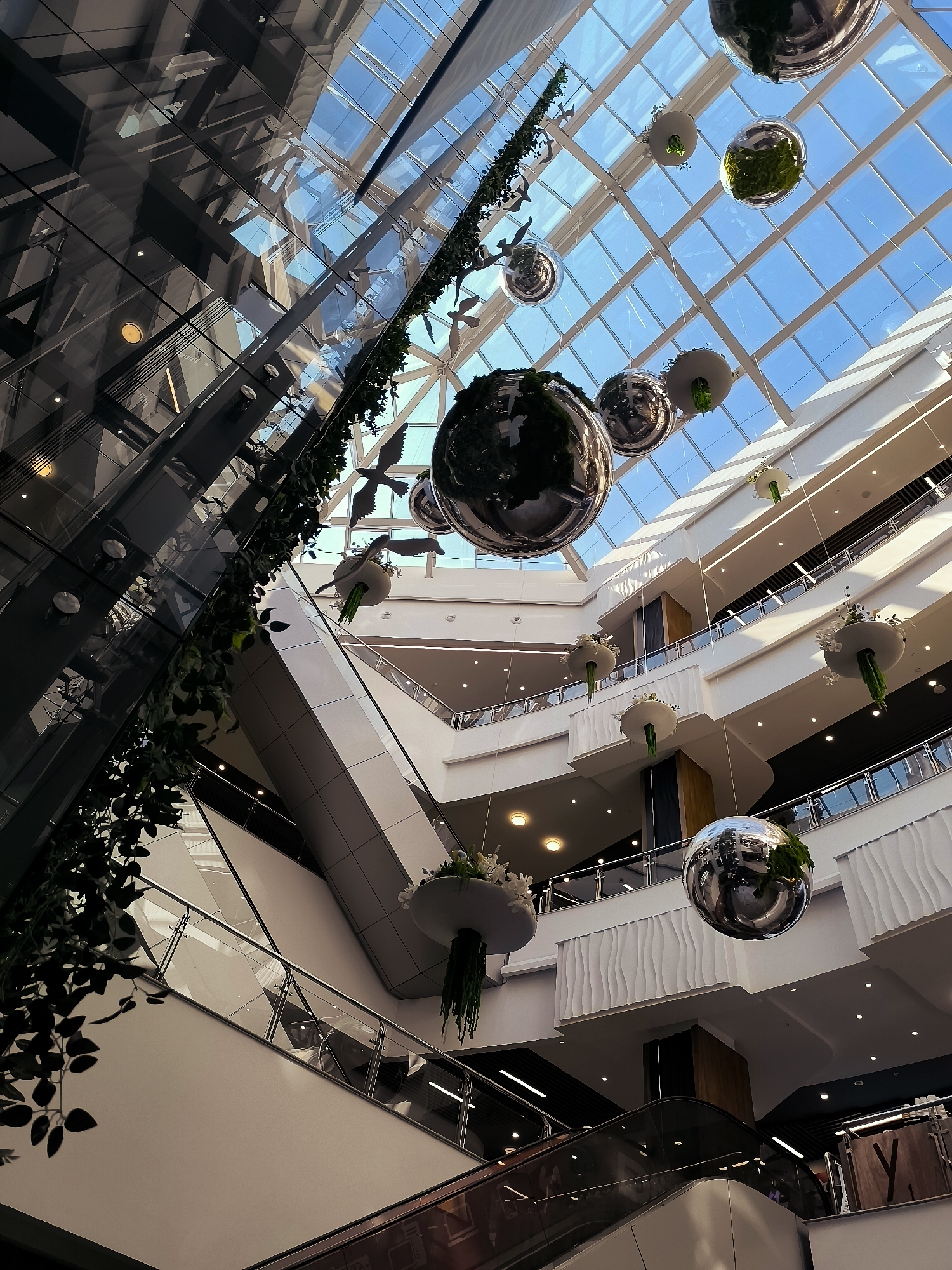 This image depicts the interior of a multi-level shopping mall, likely within the Qarağandı city administration building in Kazakhstan, given the prompt's context. The architecture features a prominent glass ceiling, allowing natural light to illuminate the space. Suspended from the ceiling are several large, spherical mirrored decorations, some of which appear to contain greenery. White, curved architectural elements are visible on the interior walls of the different levels. Escalators and staircases connect the various floors, with railings made of glass and metal. Lush green vines cascade down from a tall structure on the left side of the image. Several stylized, white bird-shaped decorations are also suspended in the atrium. The overall impression is one of a modern, bright, and aesthetically designed commercial space.