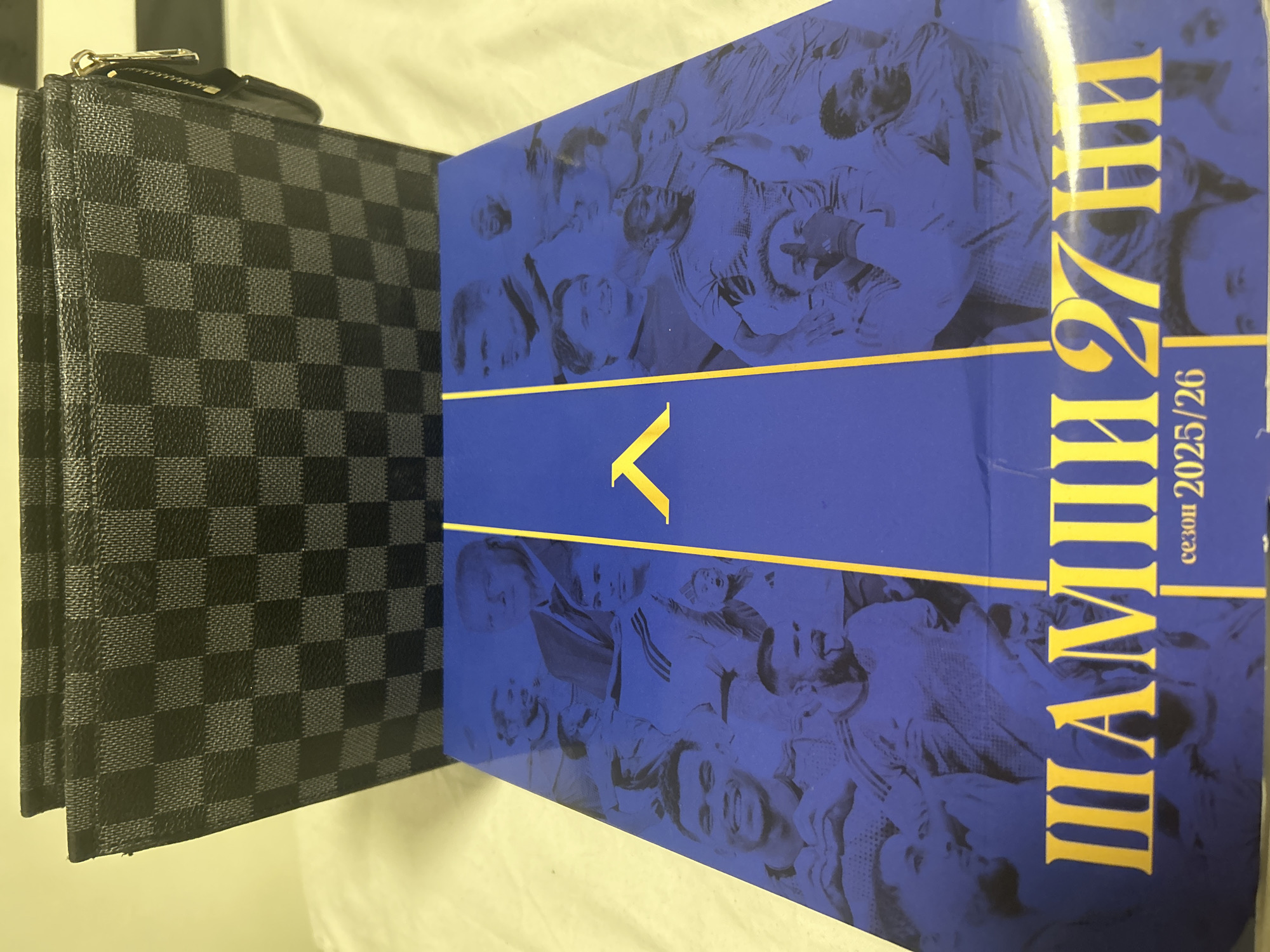 An indoor, eye-level shot captures a black and grey checkered accessory alongside a vibrant blue book or box. The accessory, resembling a Louis Vuitton Damier Graphite wallet or clutch, stands upright on the left side of the frame, showcasing its distinctive pattern and a visible zipper pull at the top.
To its right, the blue book or box is also standing upright, featuring a design with faint, halftone-style images of multiple people, possibly athletes or fans, depicted in various joyful or energetic poses. A prominent yellow horizontal stripe crosses the middle of the blue surface. Above this stripe, a stylized yellow "V" or upward-pointing arrow is visible.
Below the stripe, large yellow Cyrillic text is vertically oriented, reading "ШАМПИОНИ 27 АННИ". Directly beneath this, in smaller yellow text, is "сезон 2025/26". In Bulgarian, "ШАМПИОНИ" translates to "CHAMPIONS," and "сезон" means "season." The "АННИ" part could refer to a common feminine name in Bulgaria or another specific context.
The overall impression suggests commemorative sports merchandise, possibly celebrating a team or event related to "27" and a specific future "season 2025/26". The items are resting on a light-colored surface, likely a table, under artificial lighting. The context places the scene in Dunavtsi, Bulgaria.