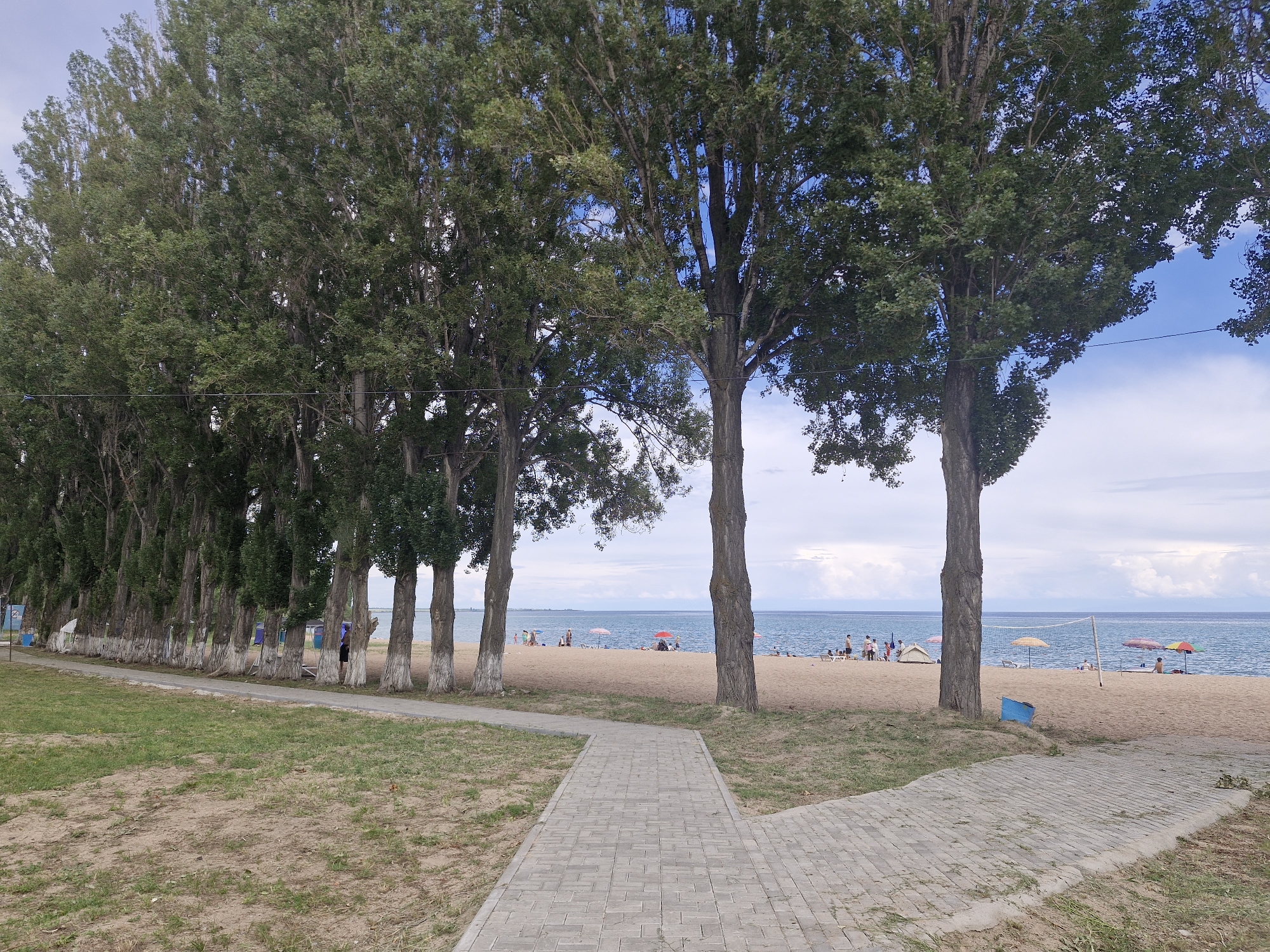 The image depicts a beach scene on the shore of Issyk-Kul Lake in Bulan-Segettu, Kyrgyzstan. A paved walkway made of grey interlocking bricks leads from the foreground towards a sandy beach. To the left of the walkway, a grassy area transitions to a line of tall, slender trees with green foliage and white-painted trunks. Beyond the trees, the sandy beach is populated with a few beach umbrellas in red, yellow, and multi-colored patterns. Several people are scattered across the beach, some standing, some sitting, and a few are near the water's edge. The water is a calm blue, extending to the horizon where it meets a sky with scattered white clouds. A volleyball net stands on the sand near the right side of the image. A blue trash bin is visible near the base of a tree on the beach. The overall atmosphere suggests a recreational day at the lake.