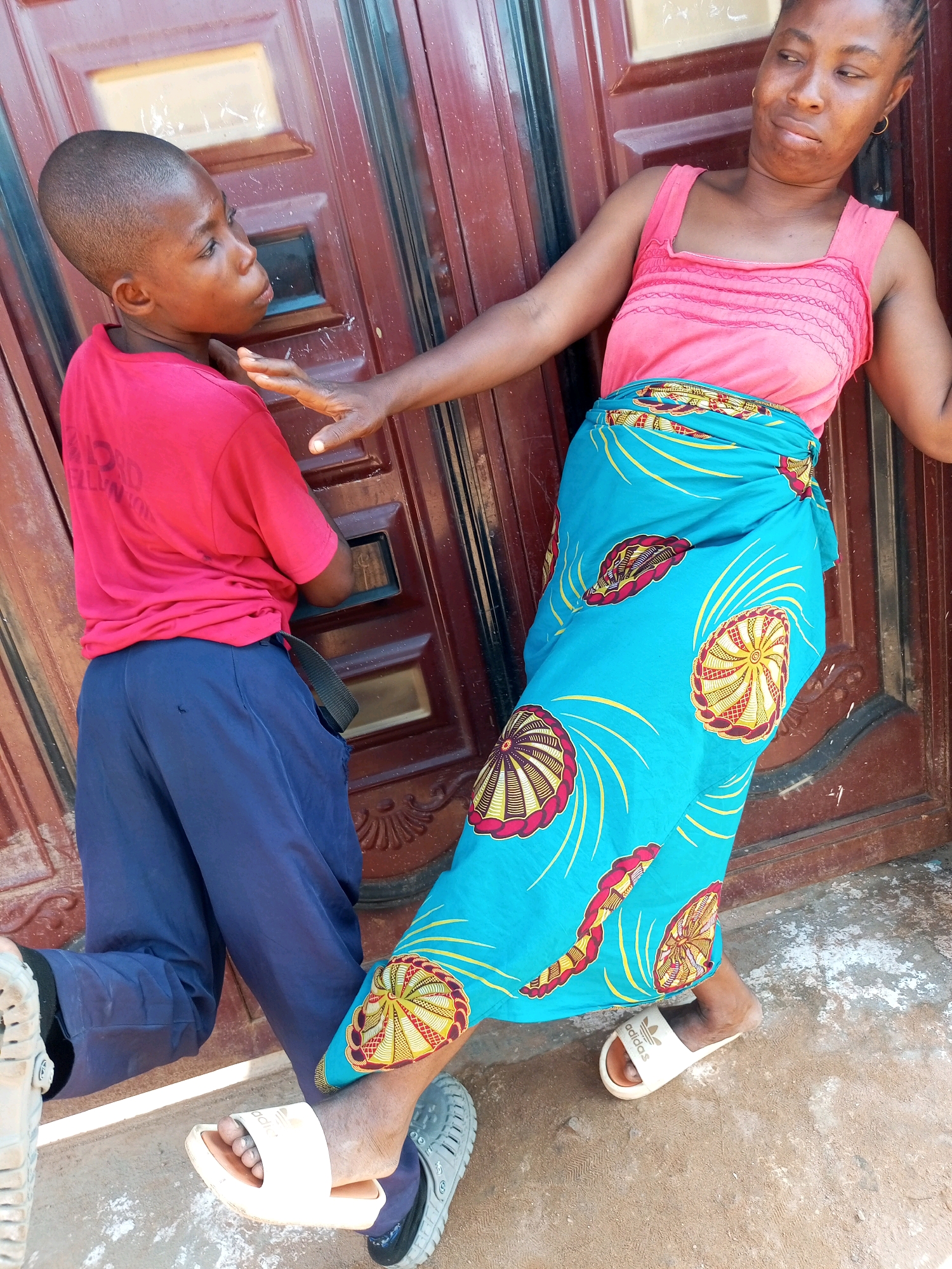 An exterior shot captures two individuals, a young boy and a woman, interacting in front of a reddish-brown wooden door in Tempure, Nigeria. The boy, on the left, faces right, wearing a bright red t-shirt and dark blue trousers. He appears to be looking away from the woman, with his left arm extended towards her. The woman, positioned on the right and facing left, wears a pink tank top and a blue wrap skirt adorned with a vibrant pattern of circular designs. Her left arm is outstretched towards the boy, with her hand resting on his shoulder or upper arm. Both individuals are wearing open-toed sandals. The ground beneath them is a dusty, textured surface. The background consists of the weathered wooden door, with its paneling and a rectangular inset that appears to be a window or a decorative element.
