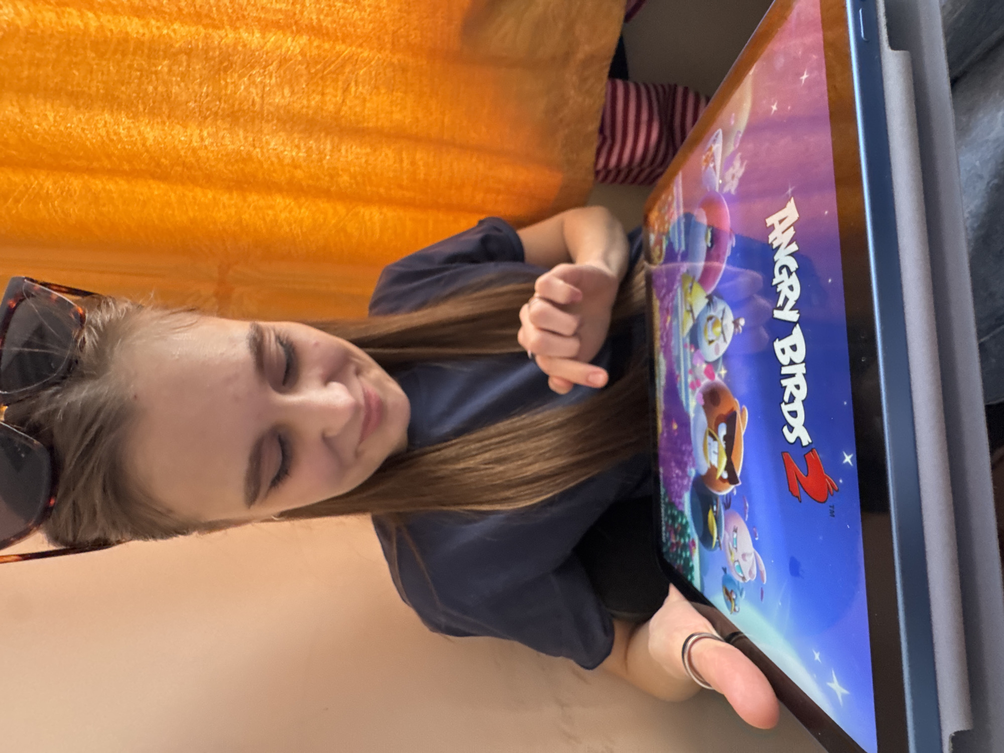 A young adult female is depicted holding and interacting with an electronic tablet in an indoor setting. The specific location within the city of Perm, Russia, cannot be determined from the visual information provided.
The female subject has long, straight, brown hair and is wearing a dark blue short-sleeved t-shirt. A pair of tortoiseshell-frame sunglasses are perched on top of her head. She displays a slight smile and is looking down at the tablet screen. A silver-colored ring is visible on her left ring finger, and a black and silver-colored ring is on her right thumb. Her right index finger is extended towards the tablet screen, indicating interaction. Her left hand supports the tablet from the side.
The tablet has a light blue bezel and is encased in a light gray protective cover. The screen displays the title screen for the video game "Angry Birds 2," featuring multiple cartoon bird characters against a blue and purple sky background.
The background consists of a plain, light peach-colored wall to the left of the subject and an orange, textured curtain or fabric to the right. A section of striped fabric, displaying pink and red horizontal stripes, is partially visible below the orange curtain on the right side of the frame.