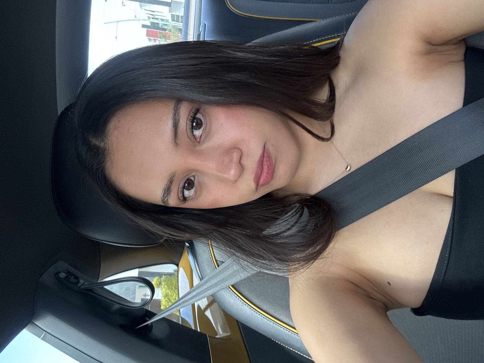 A young woman with dark hair is wearing a seatbelt while inside a car. The car's interior features black leather seats with yellow accents. The vehicle is likely parked, as the background shows buildings and trees, suggesting an urban environment. However, the precise location in Puebla, Mexico, cannot be determined from this image.