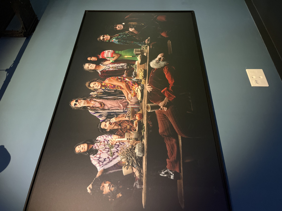 A large, rectangular, black-framed photographic print is displayed on a solid blue-green wall within The Consortium Museum (Le Consortium) in Dijon, France. The print depicts a modern reinterpretation of "The Last Supper," featuring ten individuals gathered around a long, rustic wooden table against a dark, featureless background.
Nine individuals are seated upright behind the table, arranged in a line. Their attire is varied, generally reflecting bohemian or hippie-style aesthetics with flowing fabrics, patterned garments, necklaces, and sunglasses.
From left to right:
1. A man with dark, curly hair and a beard, wearing dark clothing. His left hand is near his head.
2. A woman with long dark hair, wearing a purple and white patterned kaftan-like garment and holding a small plant branch.
3. A man with dark hair and a mustache, wearing a patterned shirt, looking down and eating from a bowl.
4. A man with long dreadlocks, a beard, and sunglasses, wearing multiple necklaces and a light-colored, patterned shirt under a robe.
5. A woman with curly hair, wearing a vibrant multi-colored patterned dress and large earrings, looking to her right.
6. A man with graying hair, wearing multiple necklaces and a reddish-brown patterned jacket, looking upwards.
7. A woman with dark hair, wearing a red and white striped top and possibly green pants, holding a lit candle.
8. A man with sunglasses, wearing a patterned green and blue jacket or shirt.
9. A man with dark hair, wearing a dark robe or jacket.
In the foreground, an older man with a long white beard is lying horizontally on a wooden bench in front of the table. He wears a red velvet-like jacket and reddish-brown patterned pants, holding a brass-colored goblet in his right hand and looking towards the viewer.
The wooden table is laden with various objects, including ceramic bowls, plates, clusters of dried herbs or flowers, candles in holders, a round cheese-like object, and an assortment of fruits such as grapes and apples.
The wall on which the print hangs is a uniform blue-green color. Reflections from an overhead light fixture are visible on the upper part of the print. A white electrical outlet with two sockets is present on the wall below the print, positioned to the right. The floor appears dark.