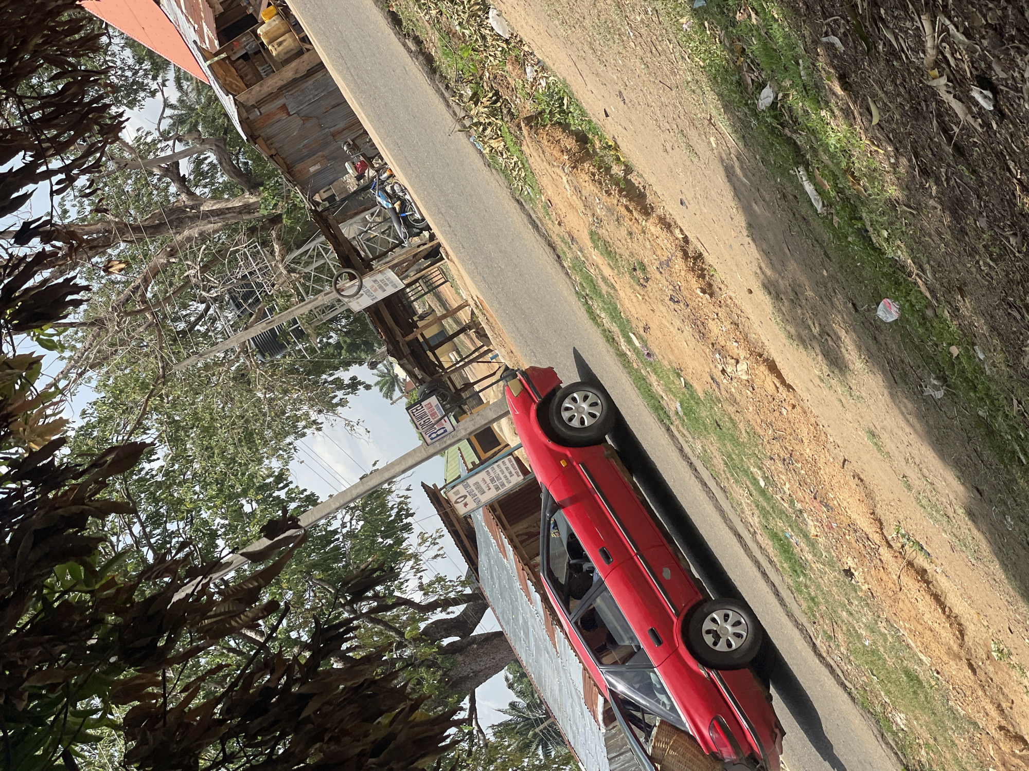 An outdoor scene captured during daytime shows a red sedan parked on the side of a paved road. The road runs diagonally through the image, separating a built-up area on the left from a dirt and grassy bank on the right.
On the left side of the road, there are several rustic buildings and structures, some featuring corrugated metal roofs and walls, indicative of local architecture in a developing region. Dense foliage and large trees frame the left side and foreground of the scene, with branches extending over parts of the buildings and the road. Utility poles with overhead wires are visible amidst the trees and structures. Behind the red car, a motorcycle or scooter is also parked.
The right side of the road consists of an unpaved, uneven dirt embankment with patches of green grass and scattered debris. The sky, visible through the gaps in the foliage, appears clear and bright, suggesting sunny weather. Shadows are cast by the car and other elements, indicating a likely morning or afternoon time of day.
Visible text on signs includes:
- A sign near the parked car reads "EKINS HOTEL," with additional, smaller text below it that is mostly illegible.
- Another sign on a pole further down the road partially reads "JESUS IS THE L" (likely "Jesus is the Lord").
- Other signs are present but too blurry to decipher.
The overall setting suggests a vibrant, potentially bustling roadside in an area like Mararaba, Nigeria, as provided in the context.