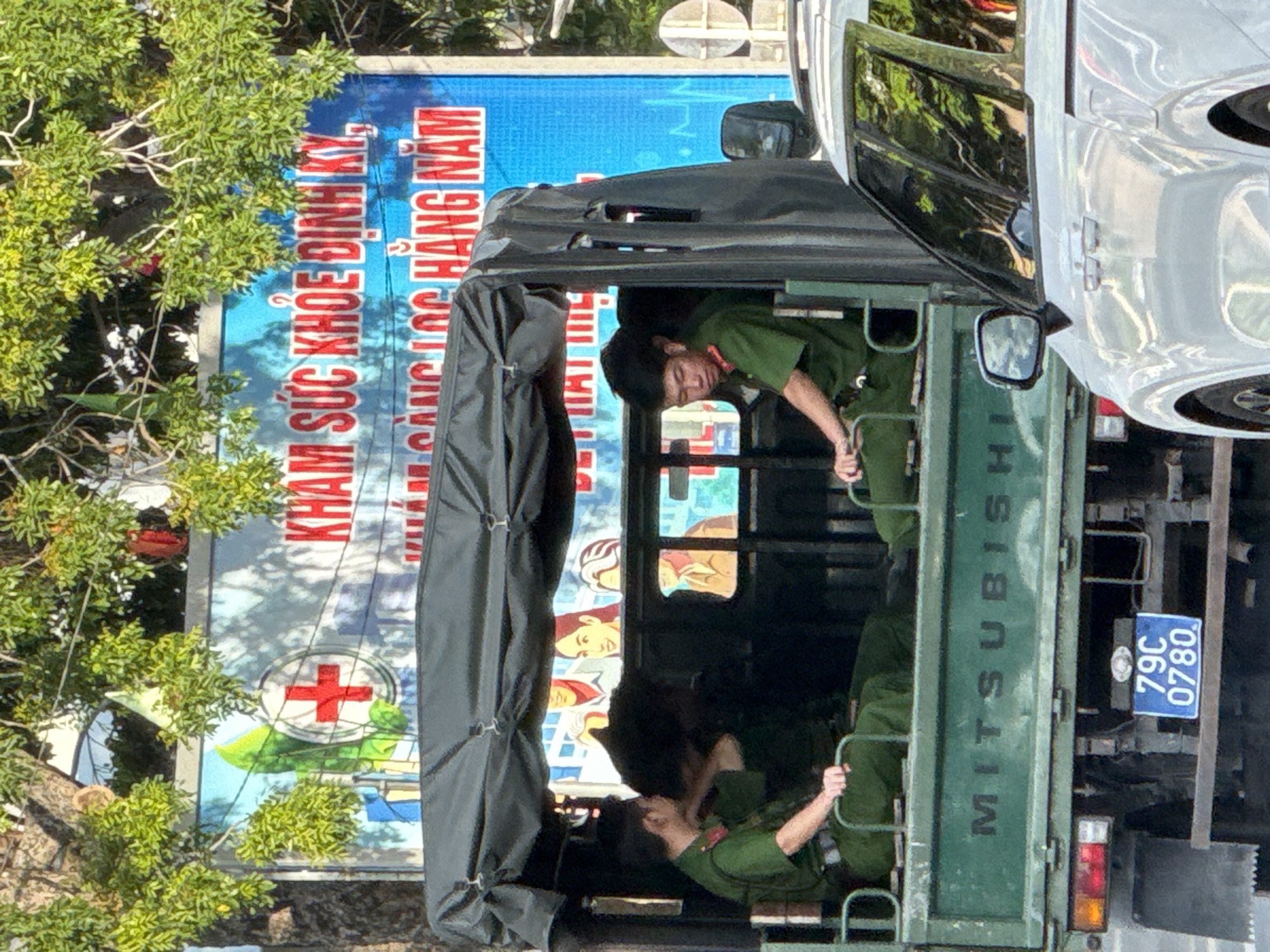 In Nha Trang, Vietnam, a green Mitsubishi truck, a utilitarian vehicle commonly used for transporting personnel, is parked. Inside the truck's open rear compartment, three individuals in green uniforms, appearing to be law enforcement or military personnel, are seated. The truck's bed is covered by a dark tarp. Behind the truck, a large blue billboard is visible, featuring a red cross emblem on a white circle, which is a common symbol for medical services, and text in Vietnamese, reading "KHAM SUC KHOE DINH KY, KHAM SANG LOC HANG NAM," which translates to "REGULAR HEALTH CHECK-UP, ANNUAL SCREENING CHECK-UP." The billboard also shows images of smiling people and medical-related graphics. The foreground is dominated by a portion of a white vehicle, suggesting other traffic on the road. Lush green foliage hangs from a tree in the upper part of the image, casting shadows. The scene appears to be a public area in Nha Trang, possibly near a clinic or hospital given the billboard's message. The license plate of the truck reads "79C 0780".