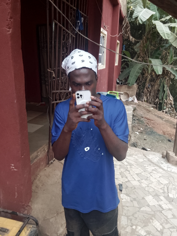 A young man in a blue t-shirt and white bandana stands outside focusing intently on his smartphone. He holds the phone with both hands, blocking his face from view. Behind him is a red building with wrought iron bars on a door or window. Vegetation including banana trees are visible behind the building. The ground has a mosaic pattern. The man is wearing dark pants. A piece of yellow equipment is visible in the lower left corner.